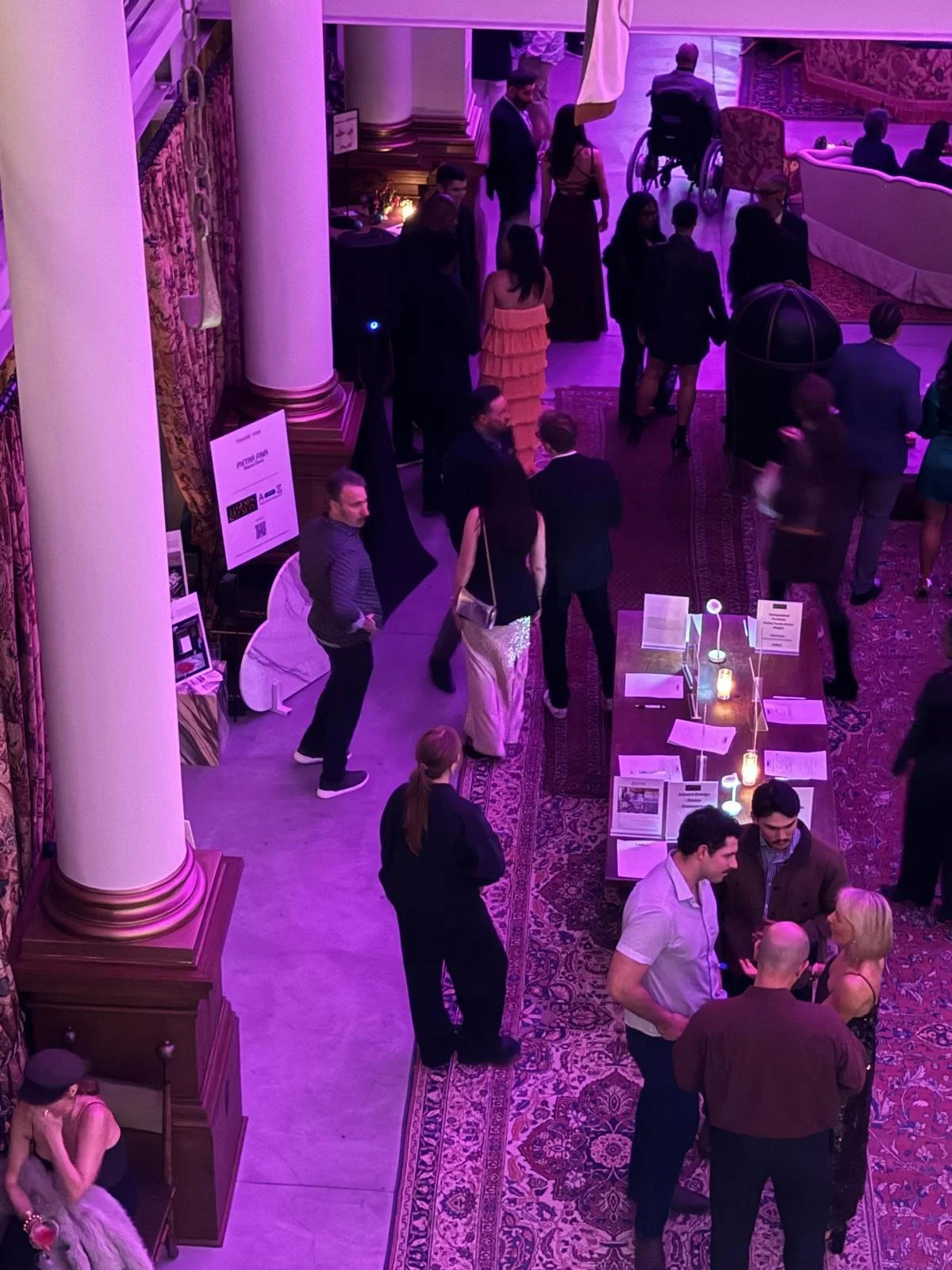 People at an elegant indoor event, some engaged in conversation, with display tables, decorative lighting, and purple ambient lighting.