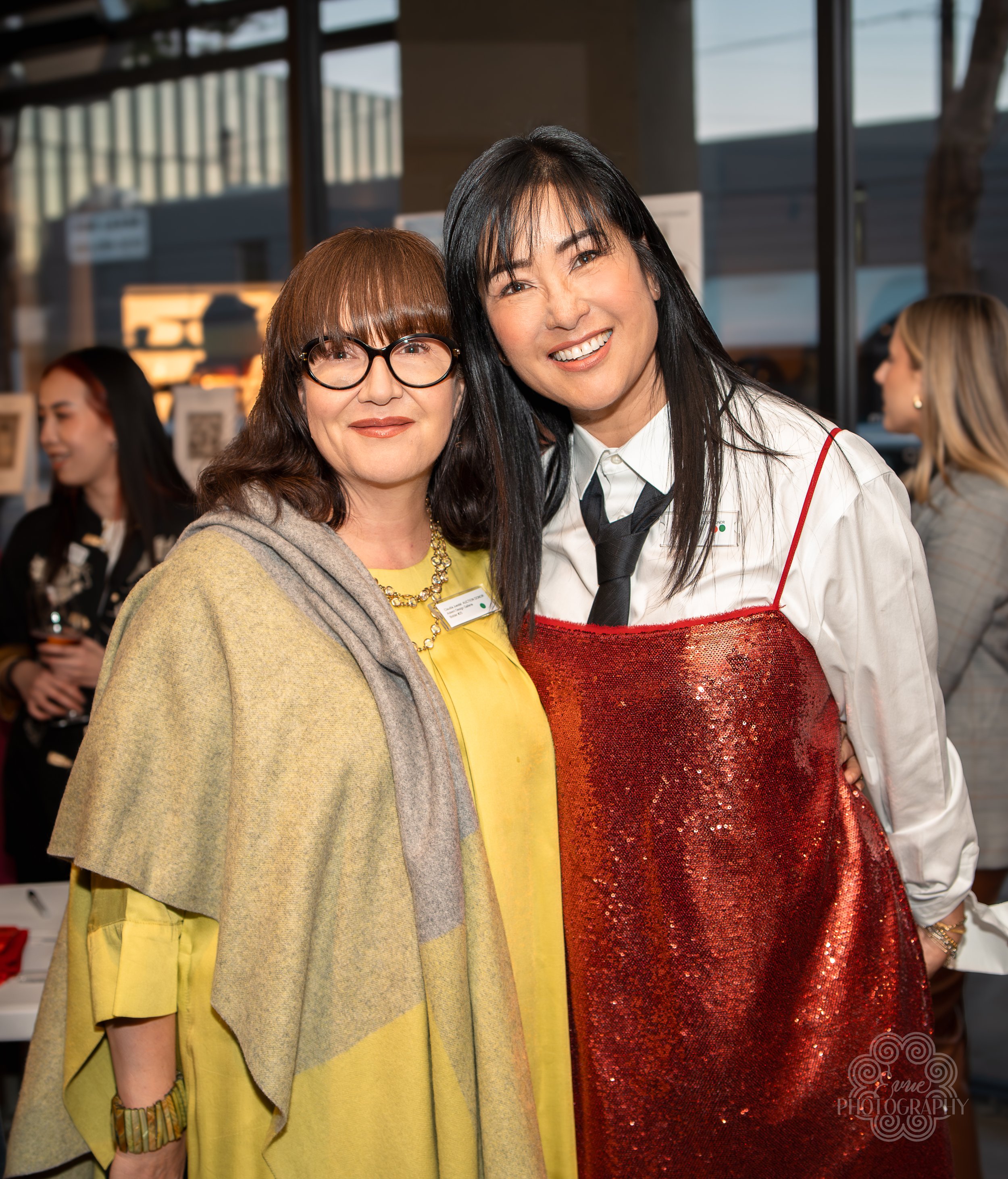 Two women standing close together indoors, smiling at the camera. The woman on the left has brown hair, glasses, and is wearing a yellow outfit with a beige and gray wrap. The woman on the right has black hair and is wearing a white shirt with a blac