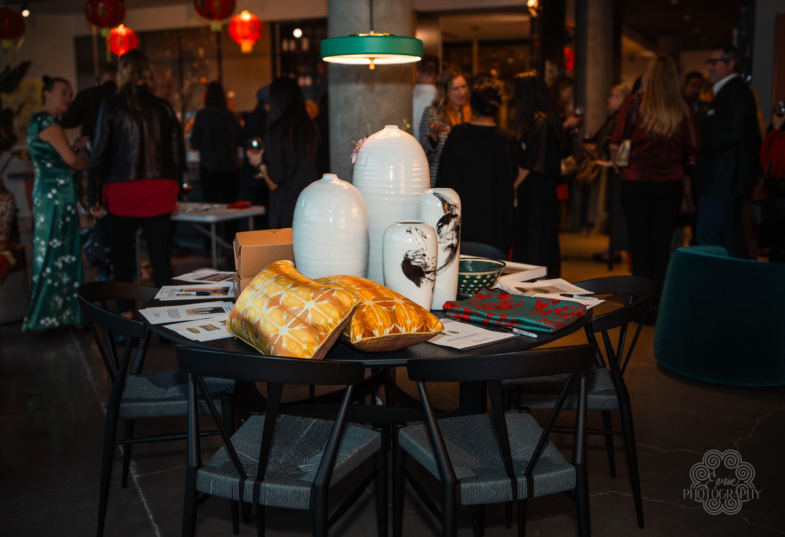 A round black table with decorative vases, pillows, and napkins, set in an indoor event space with people socializing in the background, illuminated by red lanterns.
