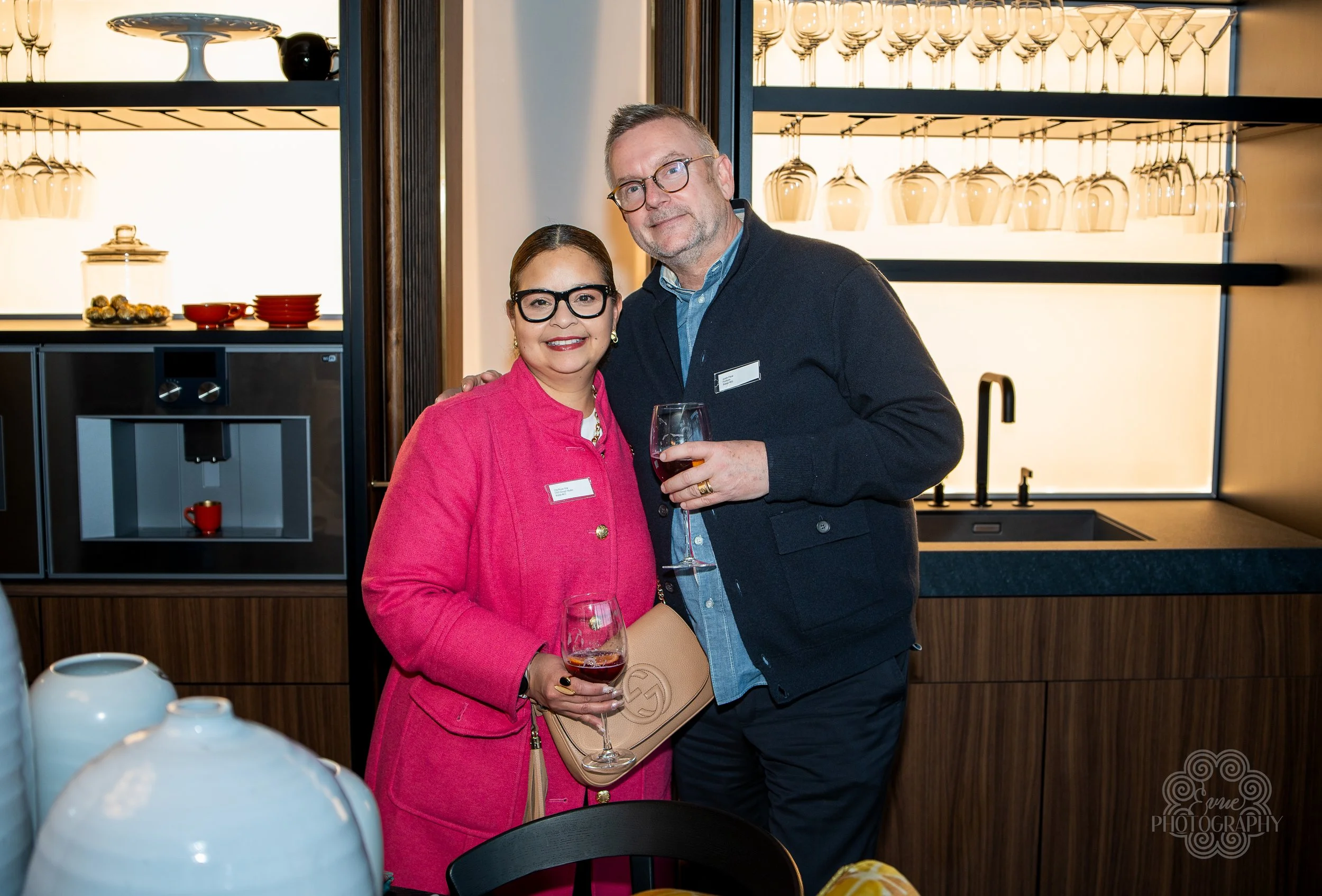 A woman and a man standing together, both holding a glass of red wine, in a modern kitchen or bar area with glassware hanging above and a sink nearby. They are smiling and posing for the photo.