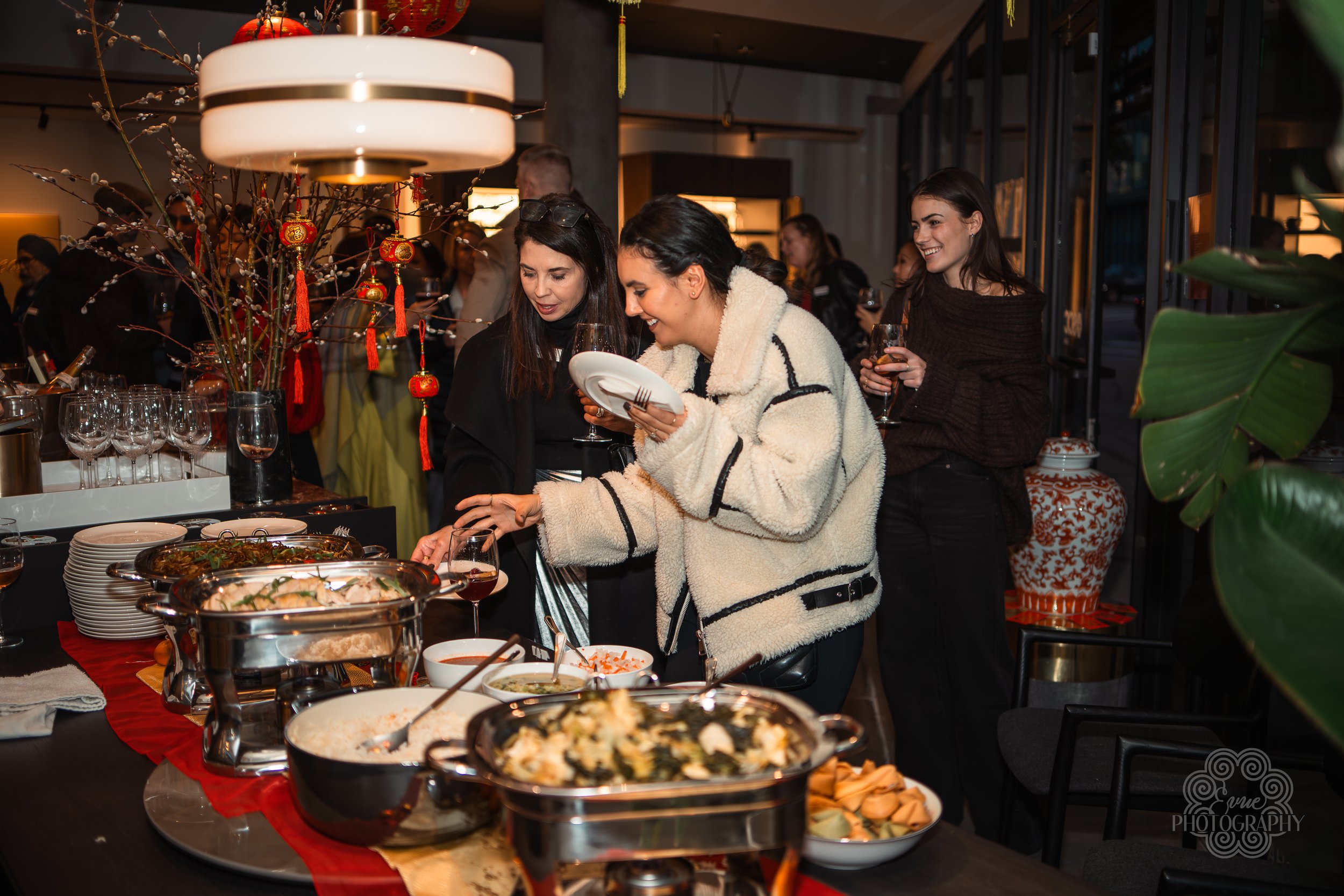 People at a buffet table enjoying a variety of Asian dishes, with decorative red and gold ornaments hanging above, in a well-lit restaurant or event space.