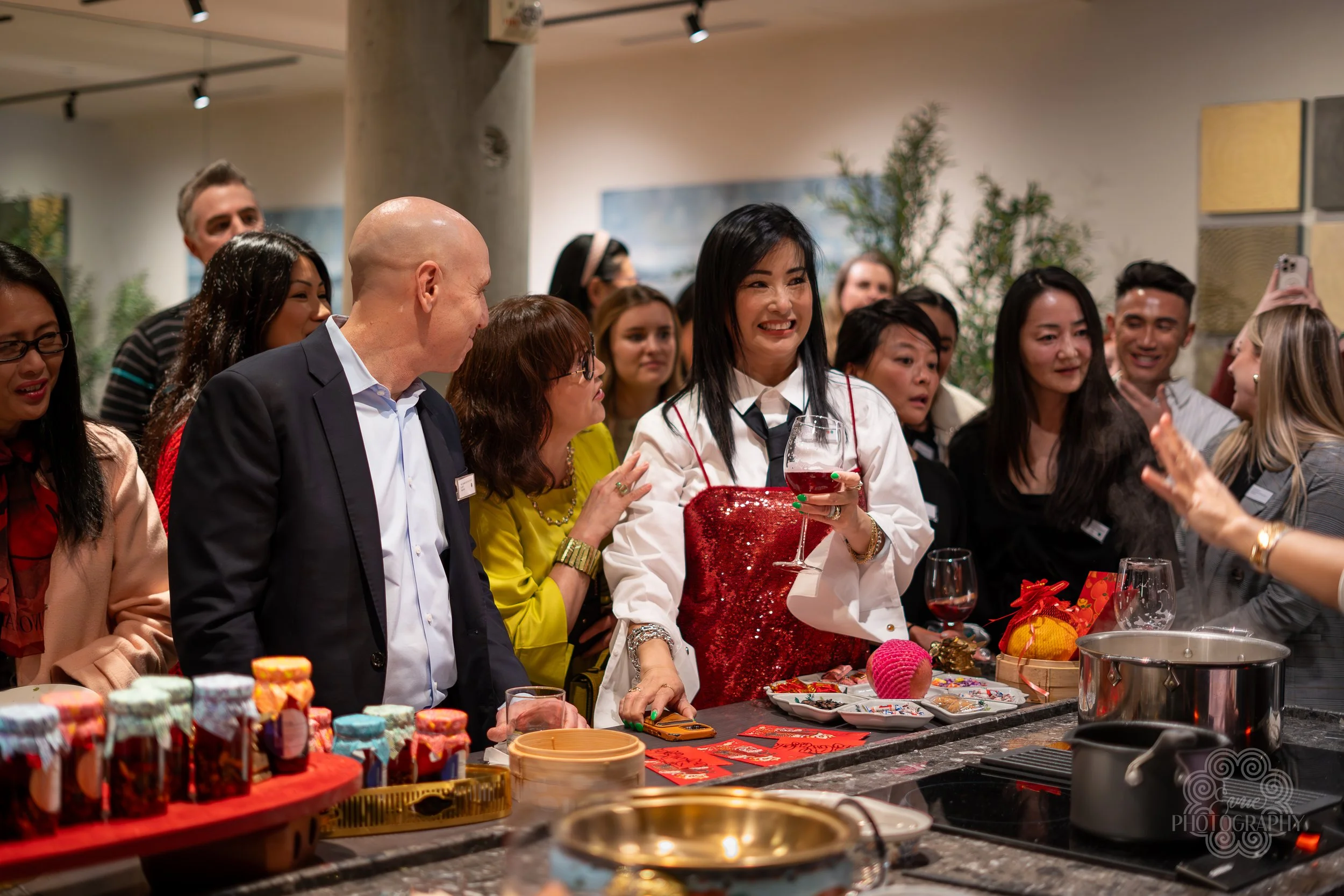 Group of people celebrating at a social gathering, with some holding wine glasses, standing around a kitchen counter with food and decorations.