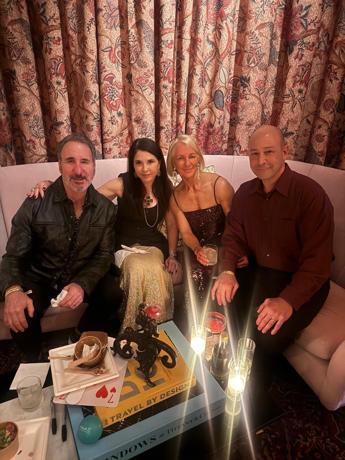 Four people sitting on a sofa at a party, with a decorative curtain behind them. There are drinks on the table in front of them, and the group appears to be enjoying a celebratory moment.