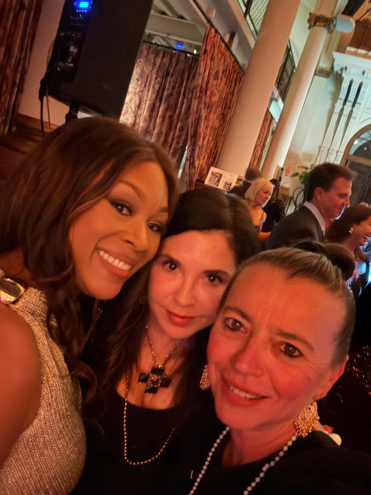 Three women taking a selfie at an indoor event, smiling, with a background of other people and decorative curtains.