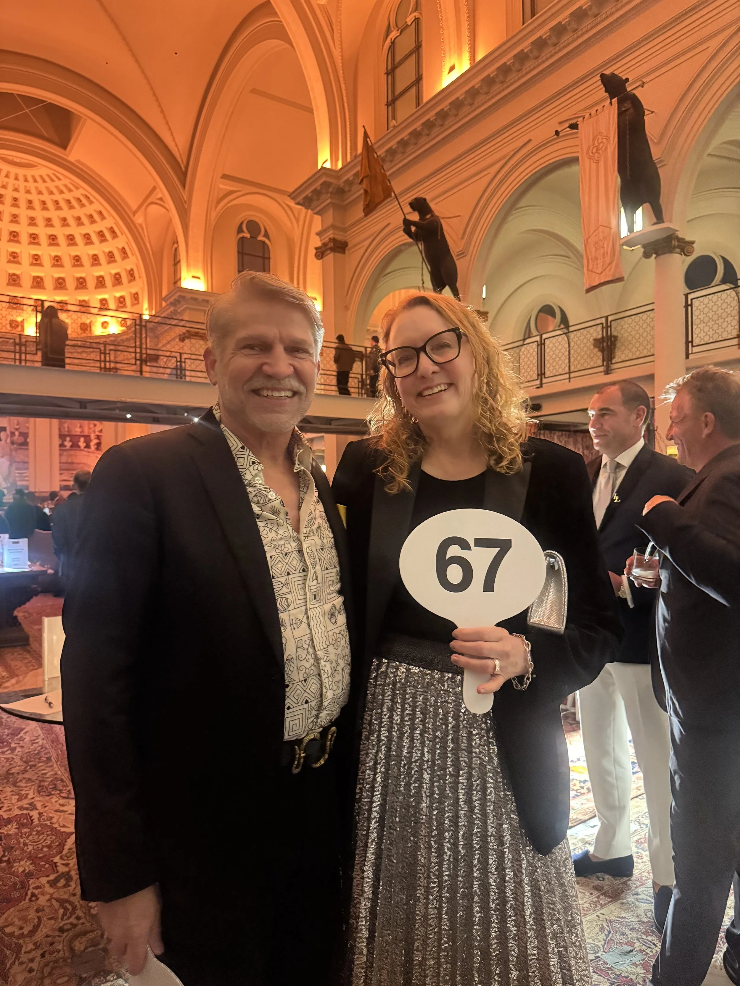 Two people smiling at an event with a grand interior and banners. The woman holds a paddle with the number 67.