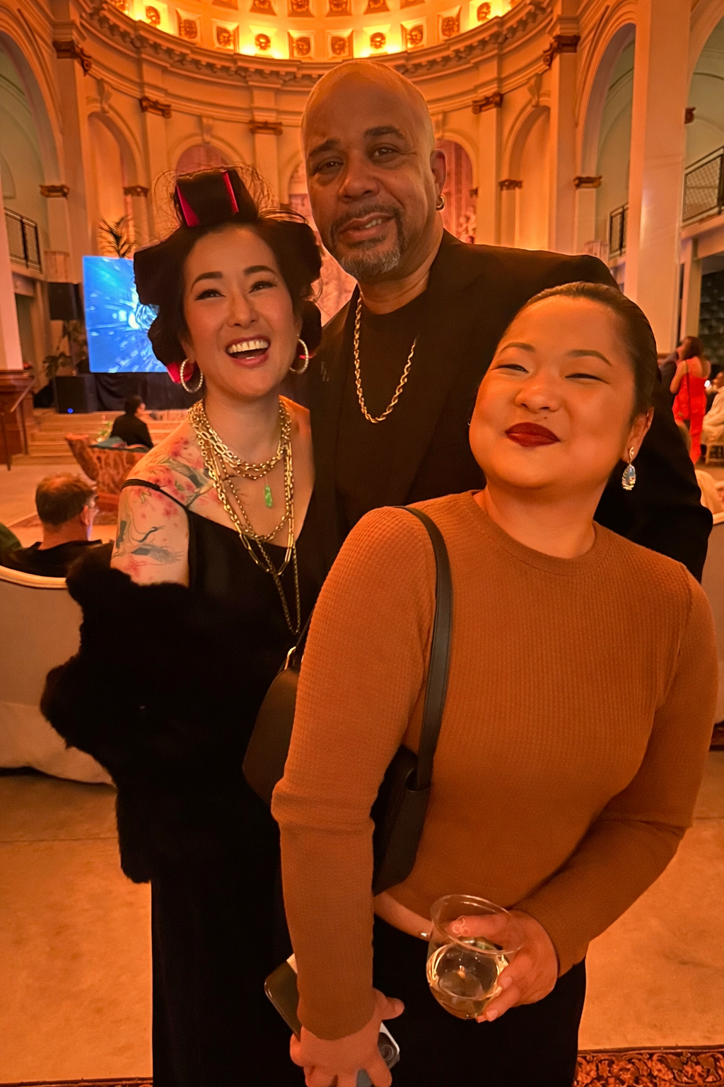 Three people posing together at an elegant event in a grand, ornately decorated hall with warm lighting and a large screen in the background.