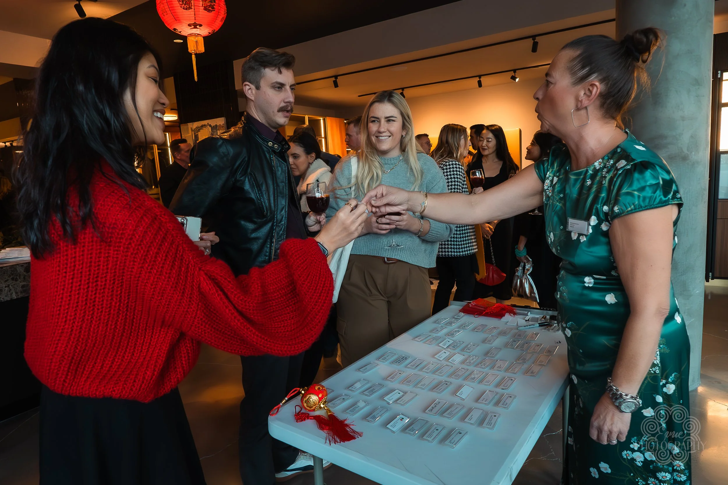 People at a social event exchanging small items, with a woman in a green dress handing something to a woman in a red sweater, while others stand around a table with name tags and decorations.