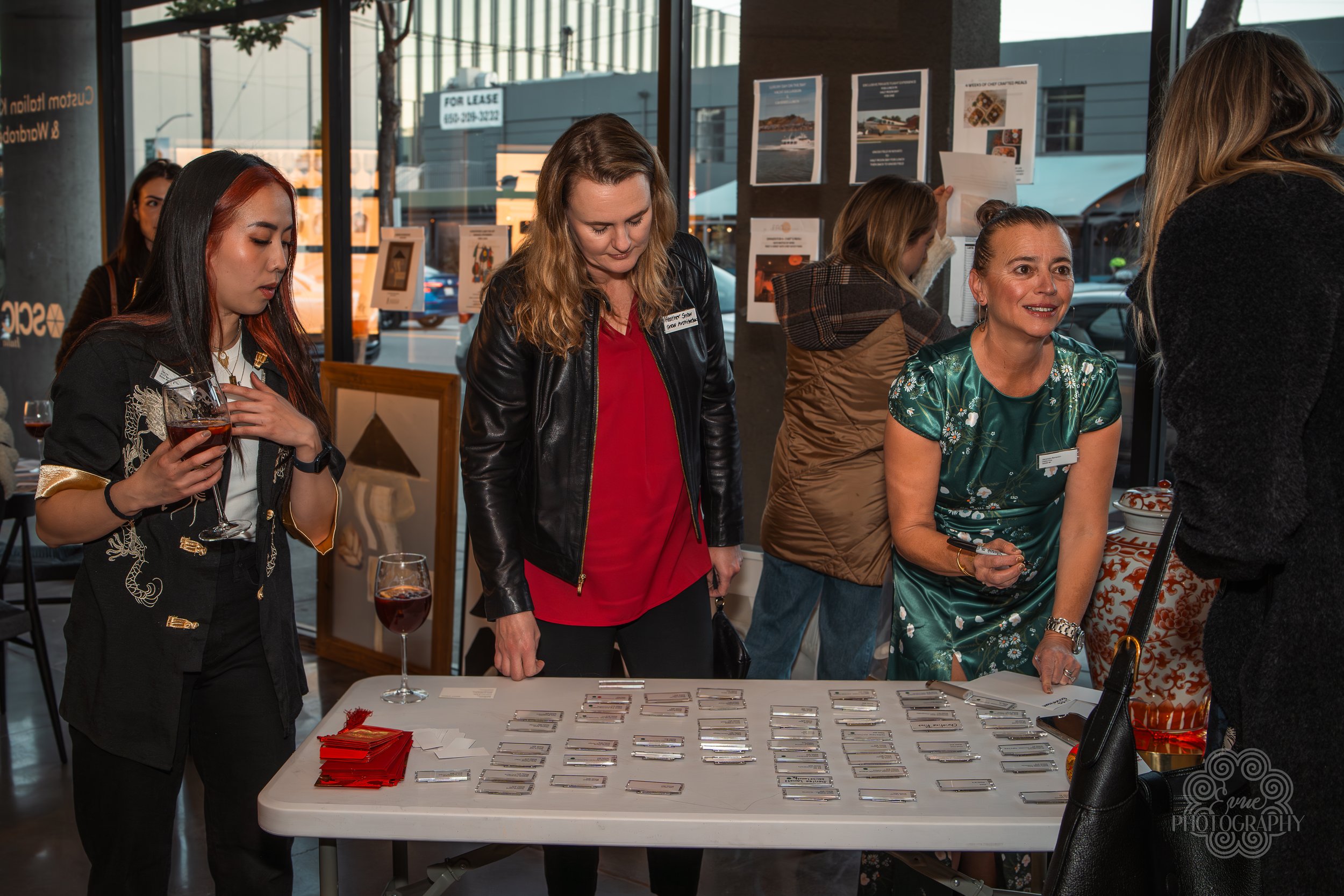 Women gathered around a table with name tags and cards at an event inside a venue with large windows and outside view of parked cars and buildings.
