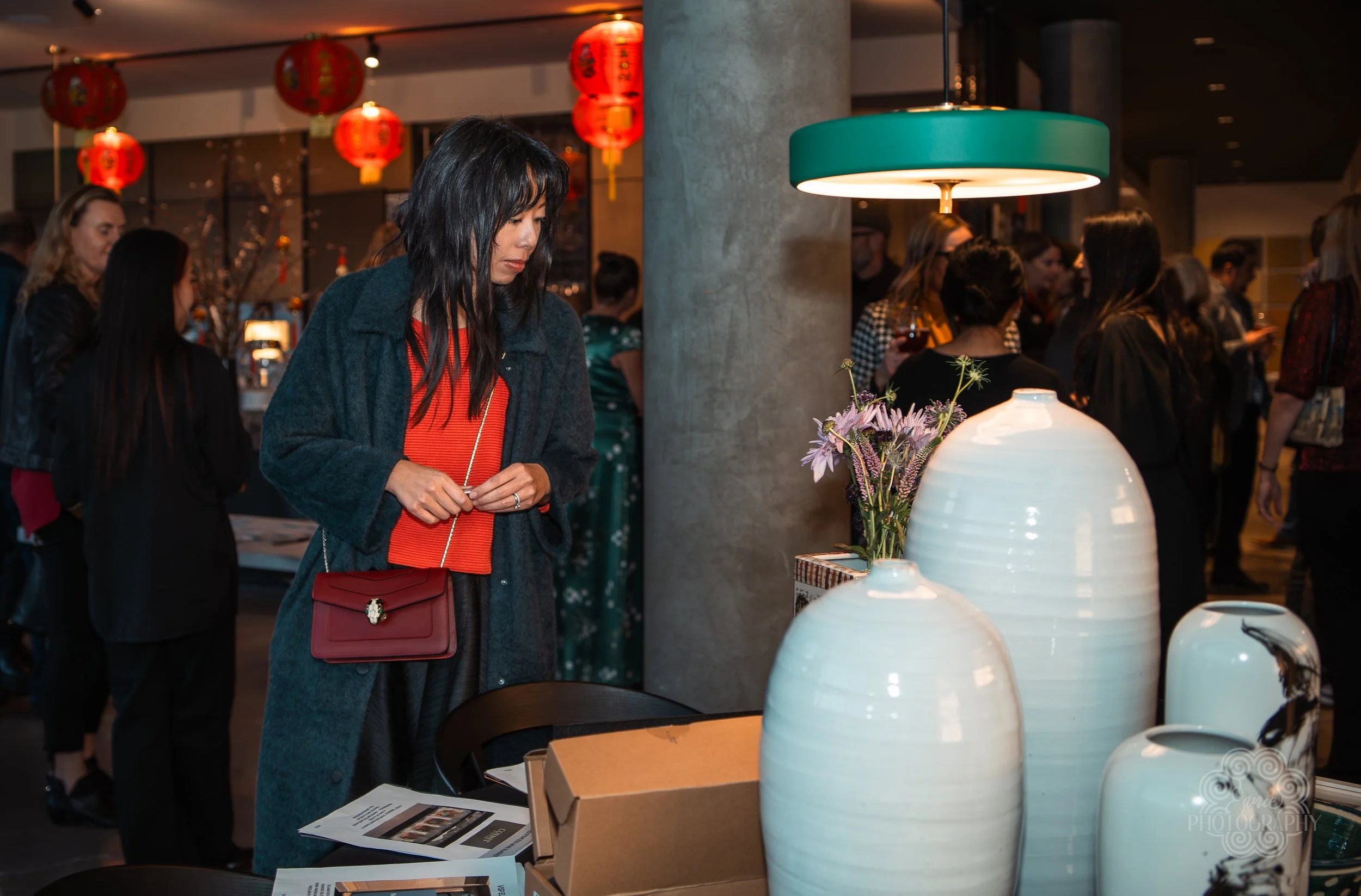 A woman with black hair, wearing a dark coat and red top, stands near decorative white vases and a flower arrangement in a busy indoor gathering space with red lanterns hanging from the ceiling.