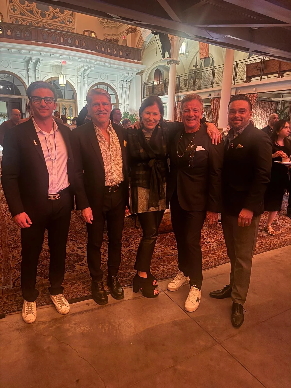 Group of five people standing together in a opulent room with elegant decor and tall columns. They are dressed in formal and semi-formal attire, smiling and posing for the photo. The background features a balcony, decorative curtains, and a vibrant s