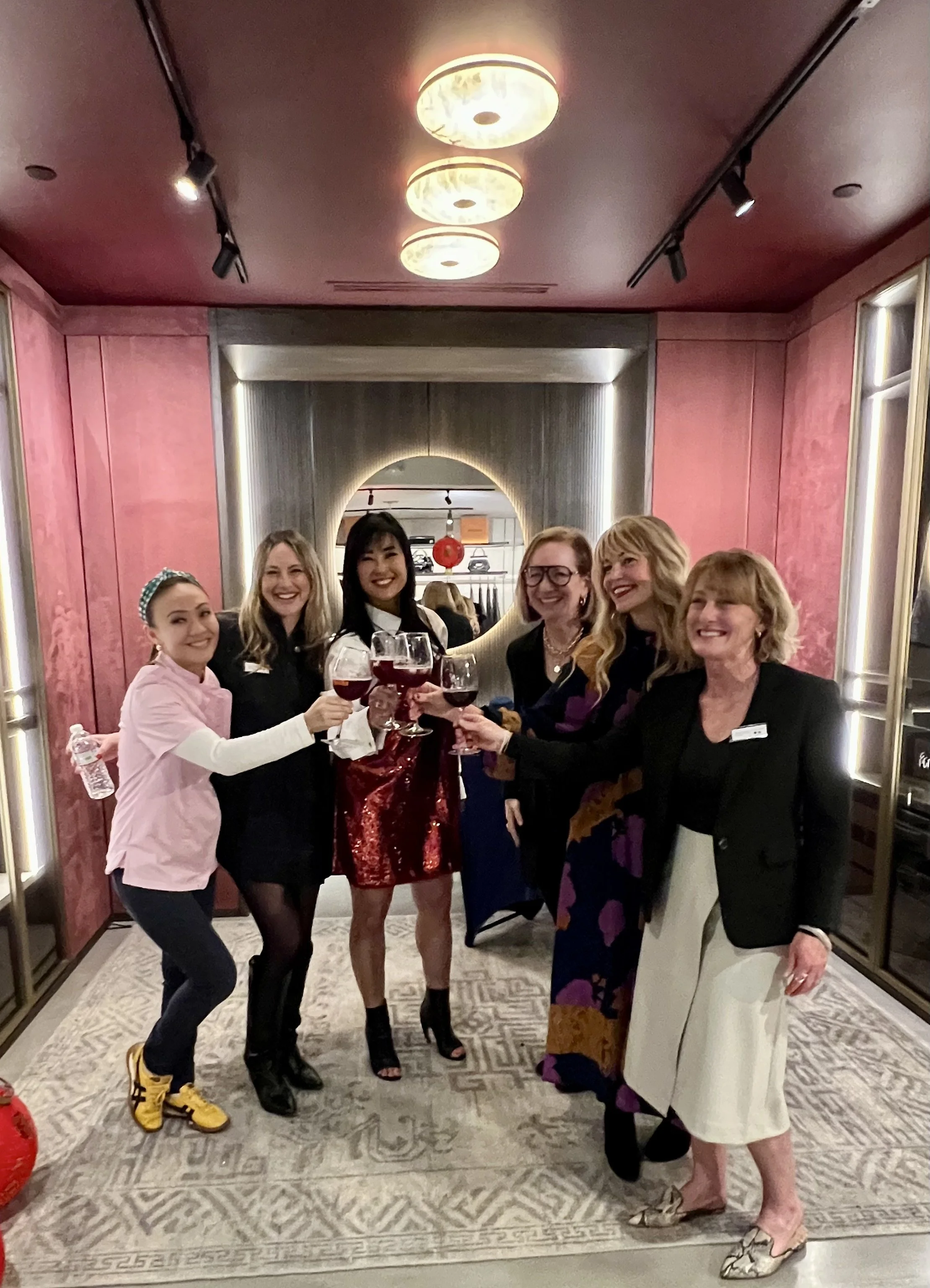 A group of six women, smiling and clinking glasses of red wine, celebrating together indoors with a pink and gray room background.