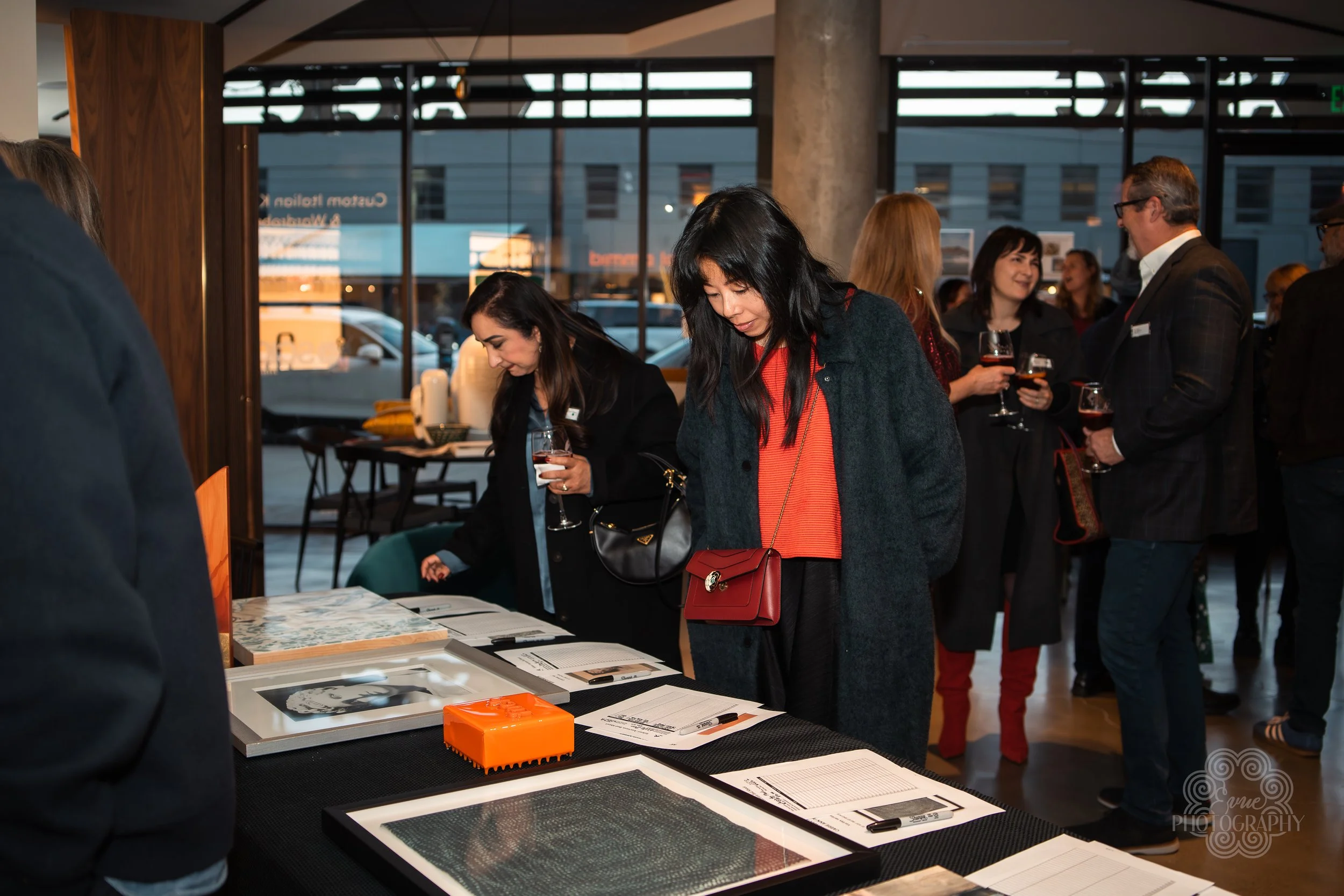 People browsing informational display tables at an indoor event, with some holding glasses of wine, in a venue with large windows and modern decor.