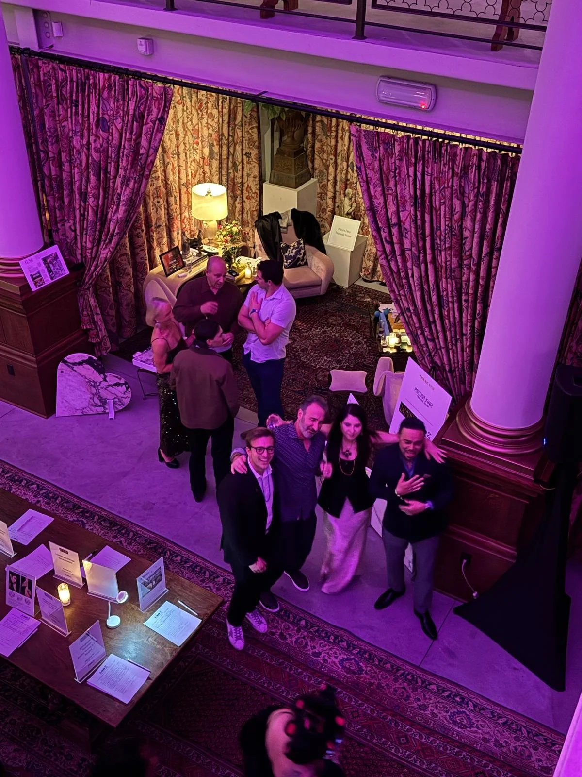 A group of people at a social event, standing and posing for a photo, inside a decorated room with curtains and table signs.