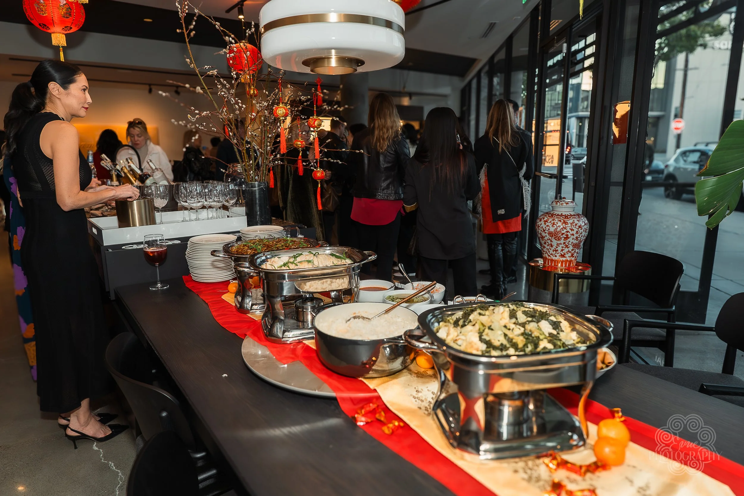 Indoor buffet setup at a restaurant with various dishes, glasses, and decorations. Guests are waiting in line near large windows.