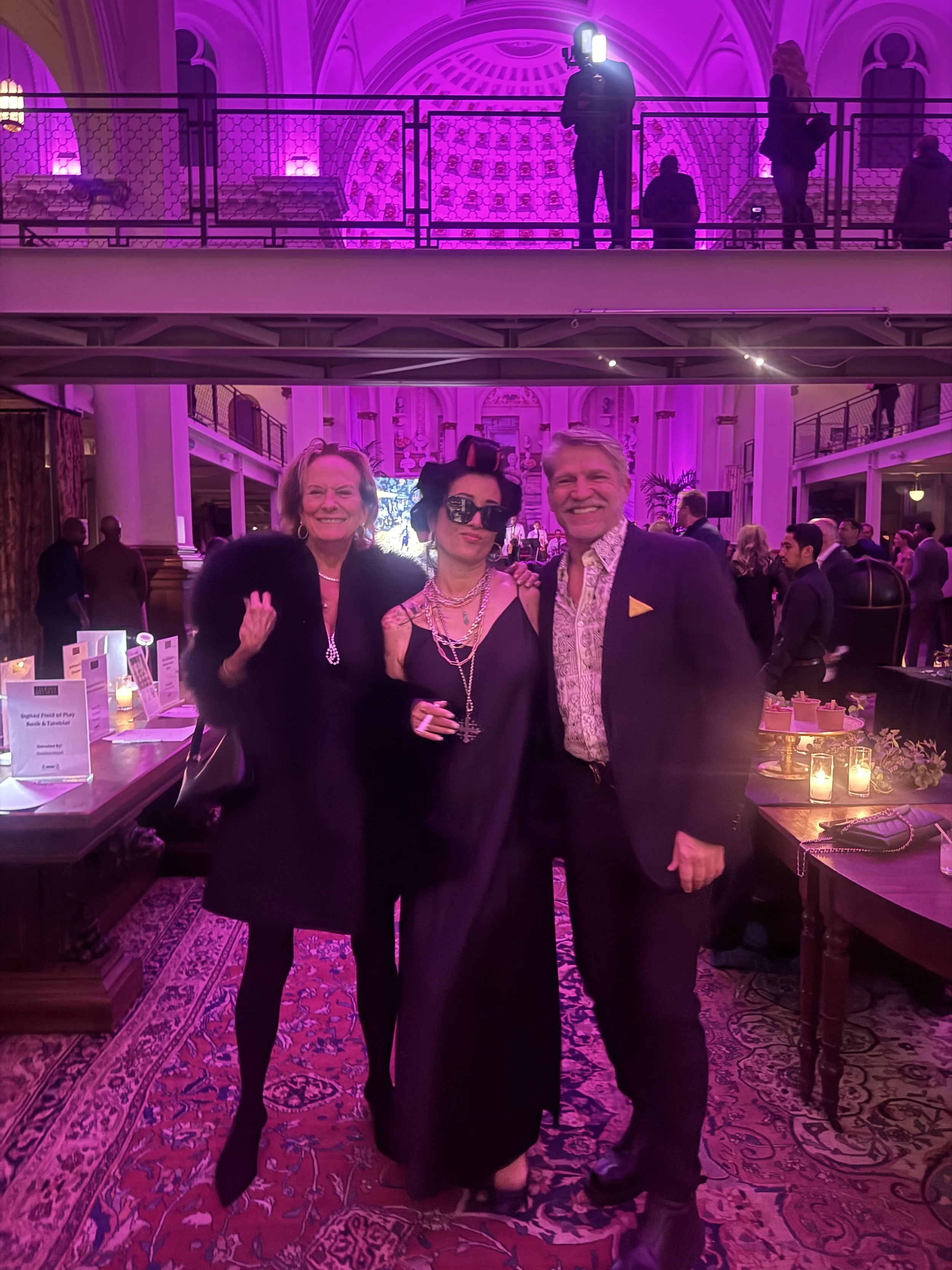 Three people posing for a photo at an indoor event with pink lighting, with people in the background and a balcony above.