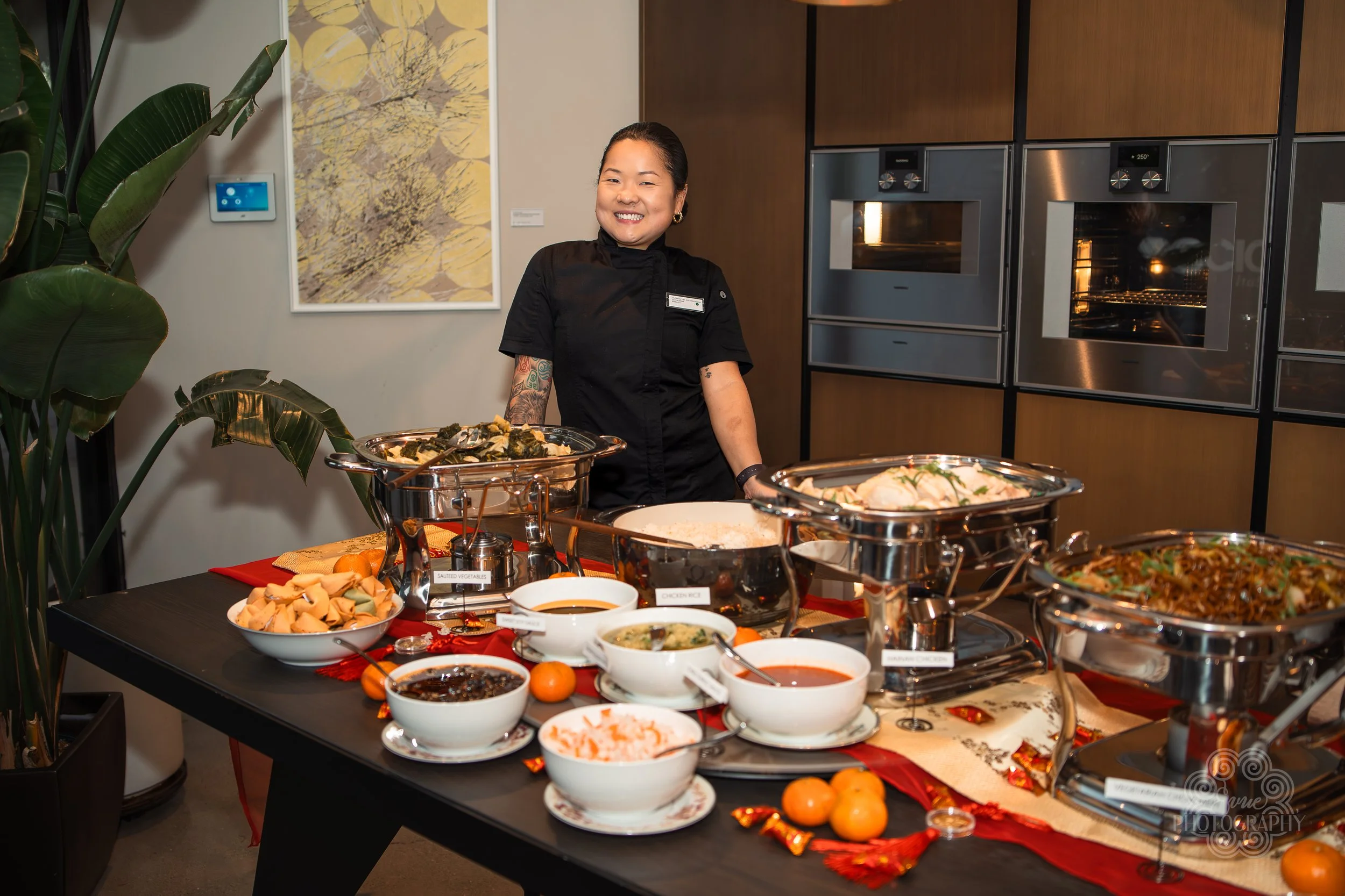 A smiling woman dressed in black standing behind a buffet table with various dishes, including soups and salads, in a modern indoor setting with potted plant and artwork on the wall.