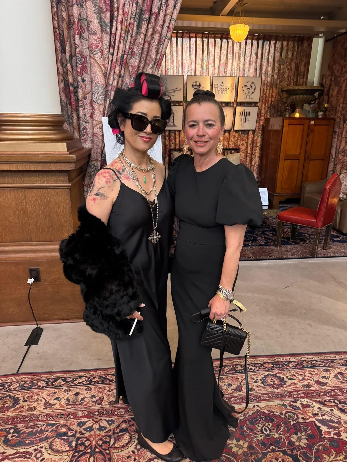 Two women dressed in black, posing together at an indoor event with patterned curtains and artwork in the background.