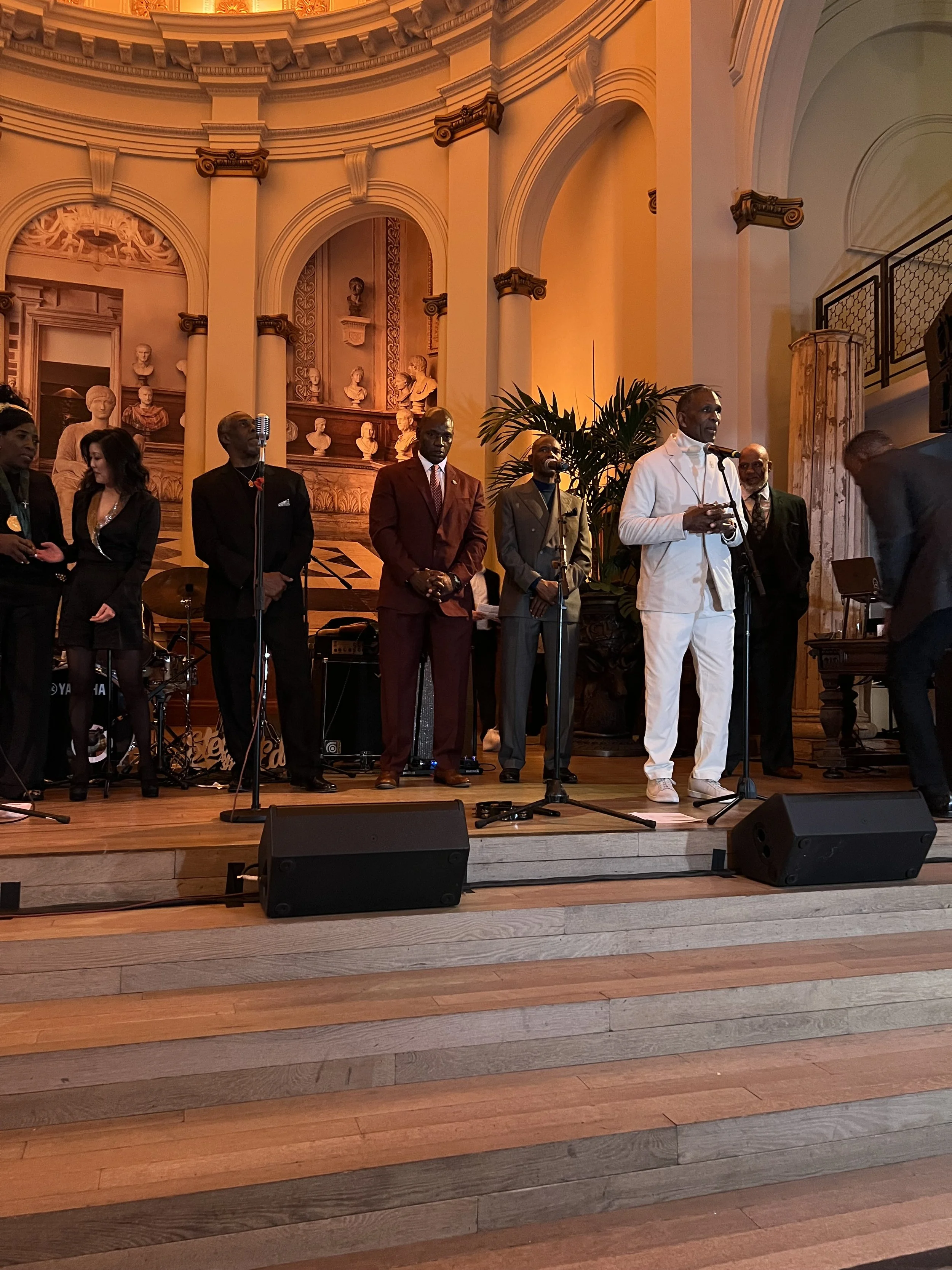 A group of men and women standing on a stage in a grand, ornately decorated hall with bust sculptures and classical architecture. One man in white is speaking into a microphone, with others dressed formally standing nearby.