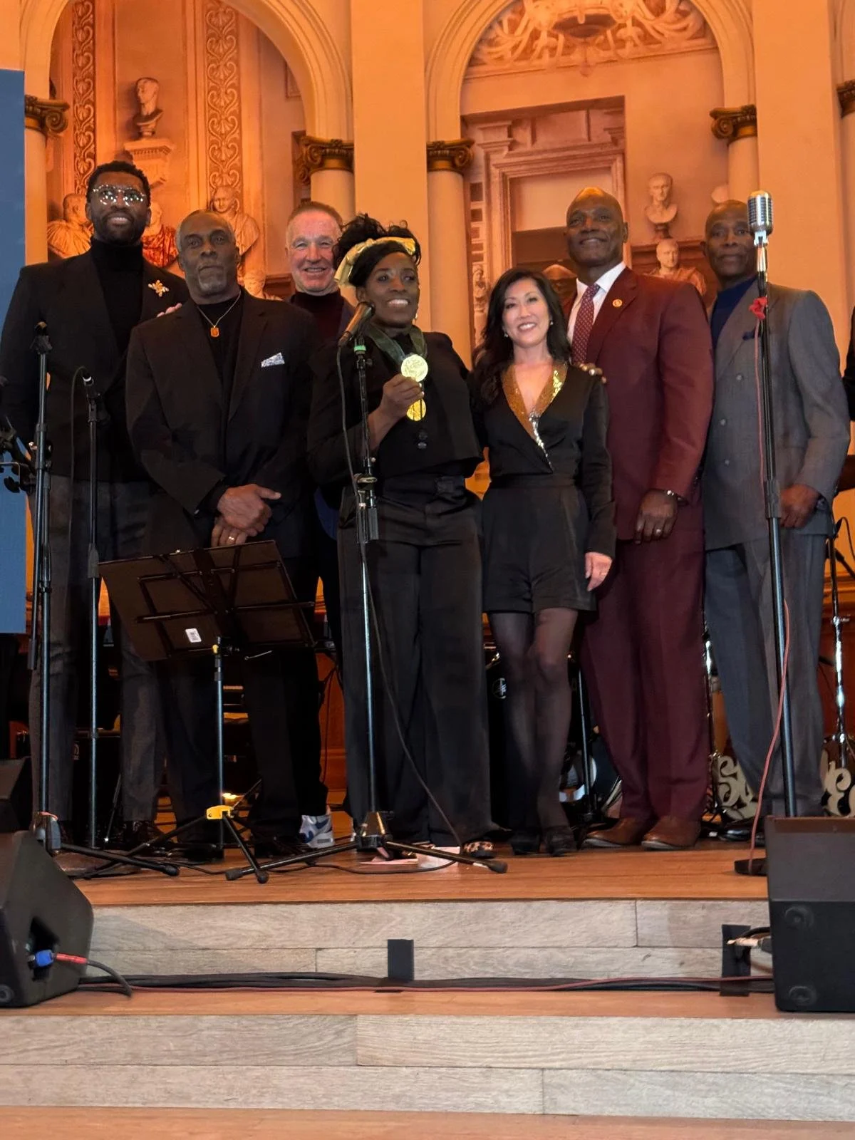 A group of eight people standing on a stage in a decorated hall, some holding microphones and medals, smiling for a photo.