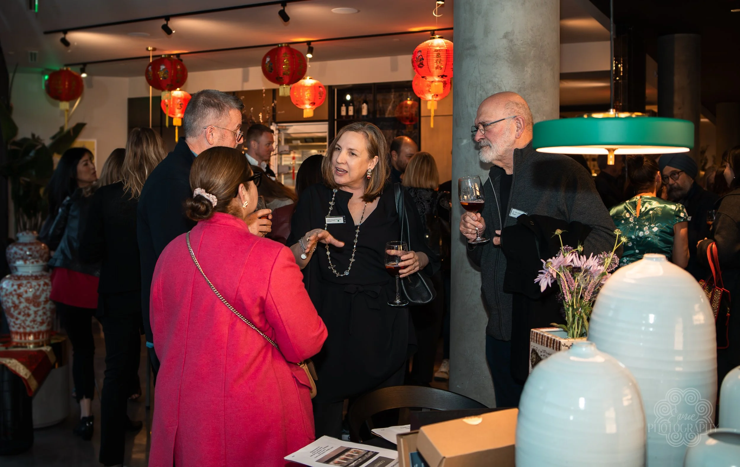 Group of people at a social gathering, holding glasses of wine and engaged in conversation inside a decorated venue with red Chinese lanterns hanging from the ceiling.