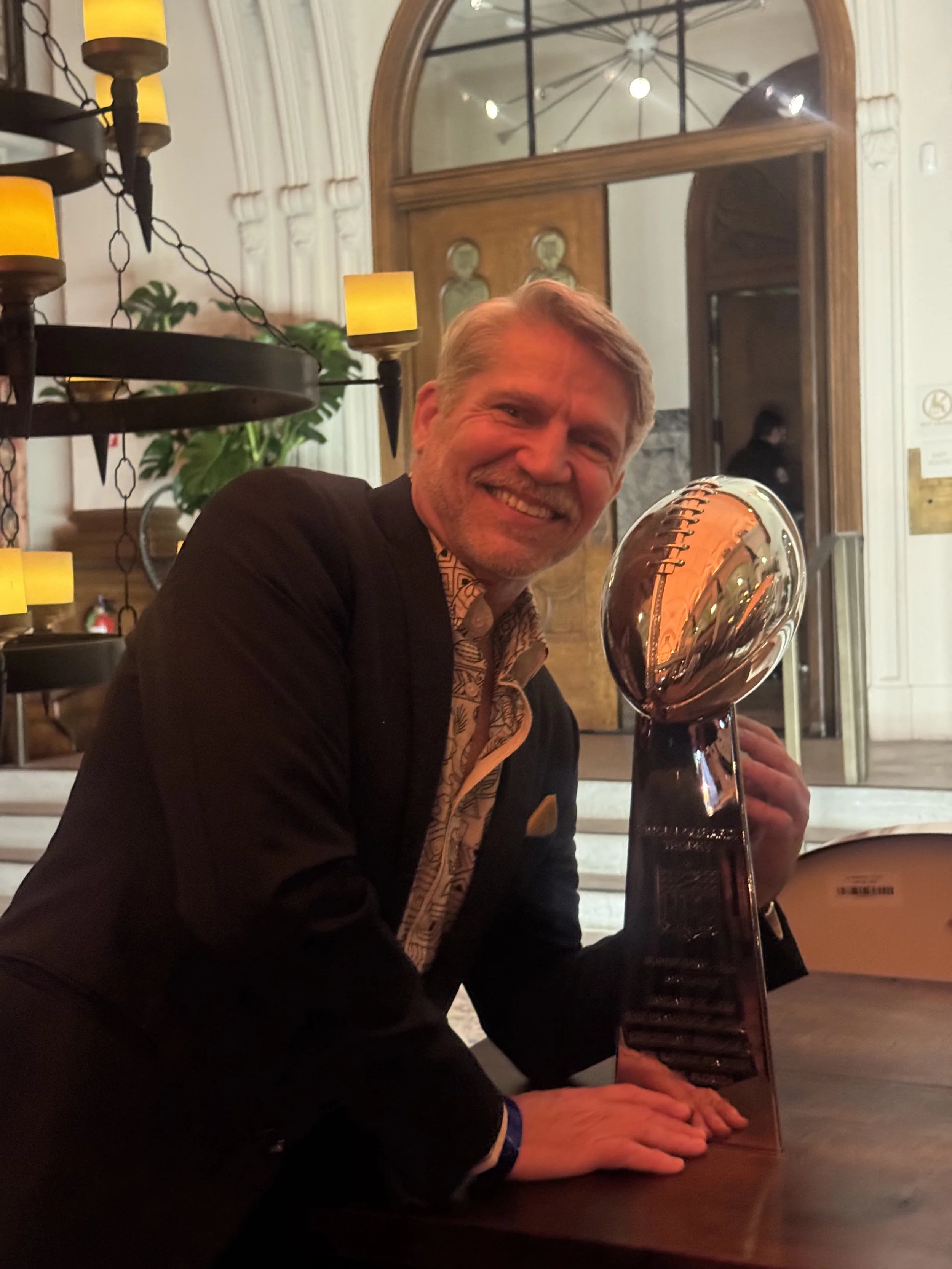 A smiling man in a dark suit holding a trophy with a shiny, metallic sphere on top in an elegant indoor setting with warm lighting and a large mirror in the background.