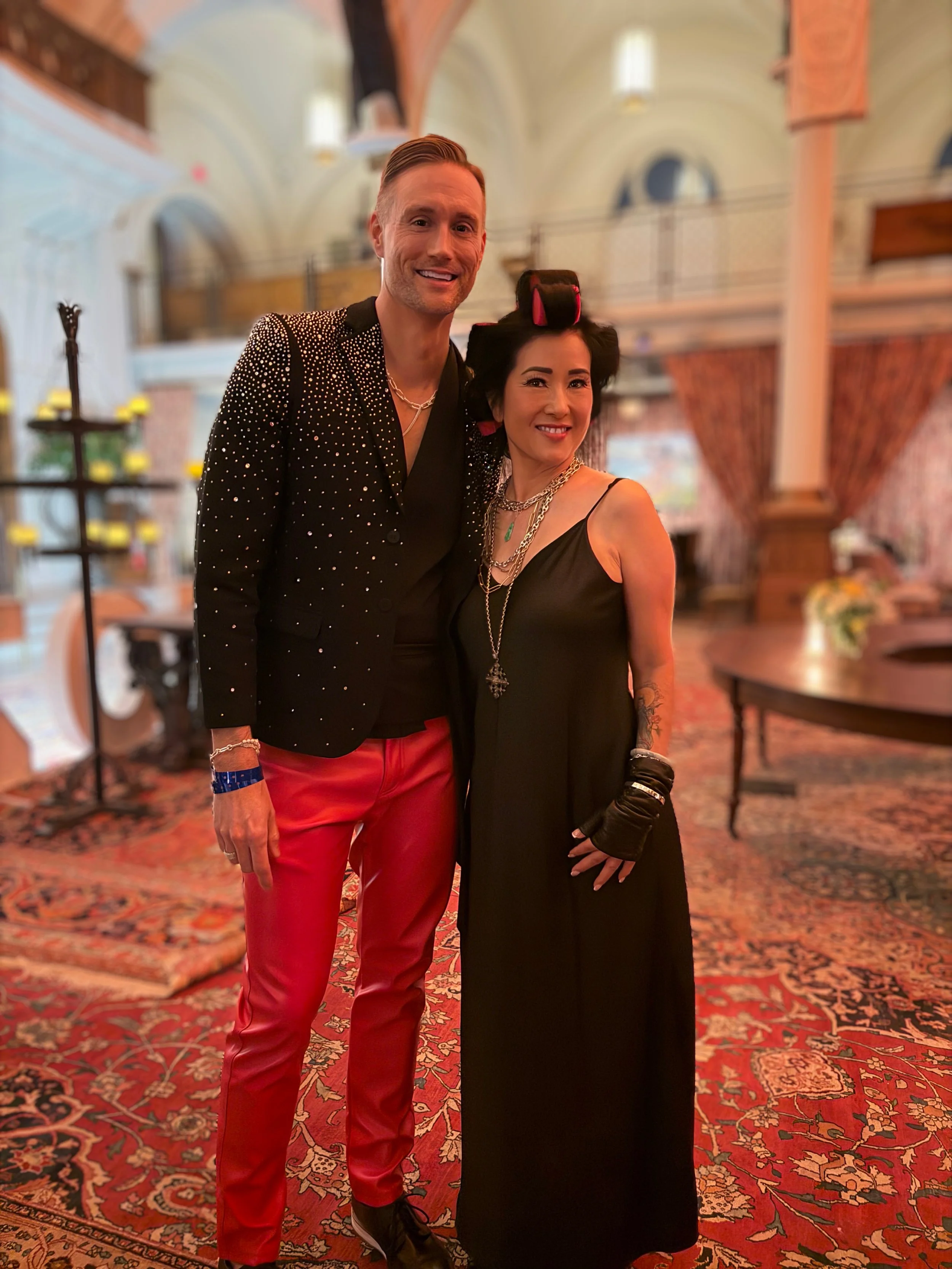 A man and a woman dressed in glamorous clothing posing together indoors with ornate décor, rugs, and curtains in the background.