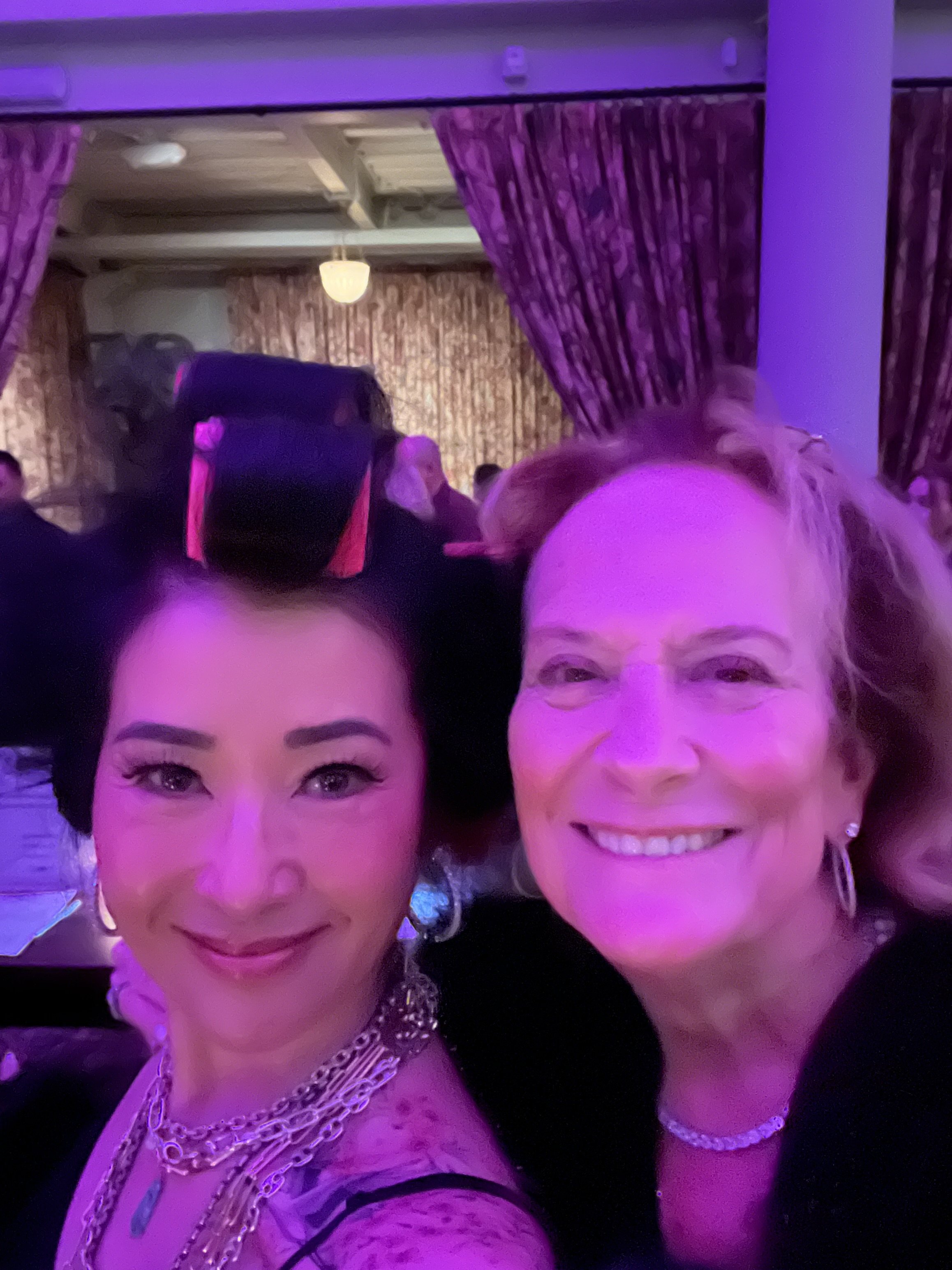 Two women taking a selfie at a social event with purple lighting, ornate curtains, and a crowd of people in the background.