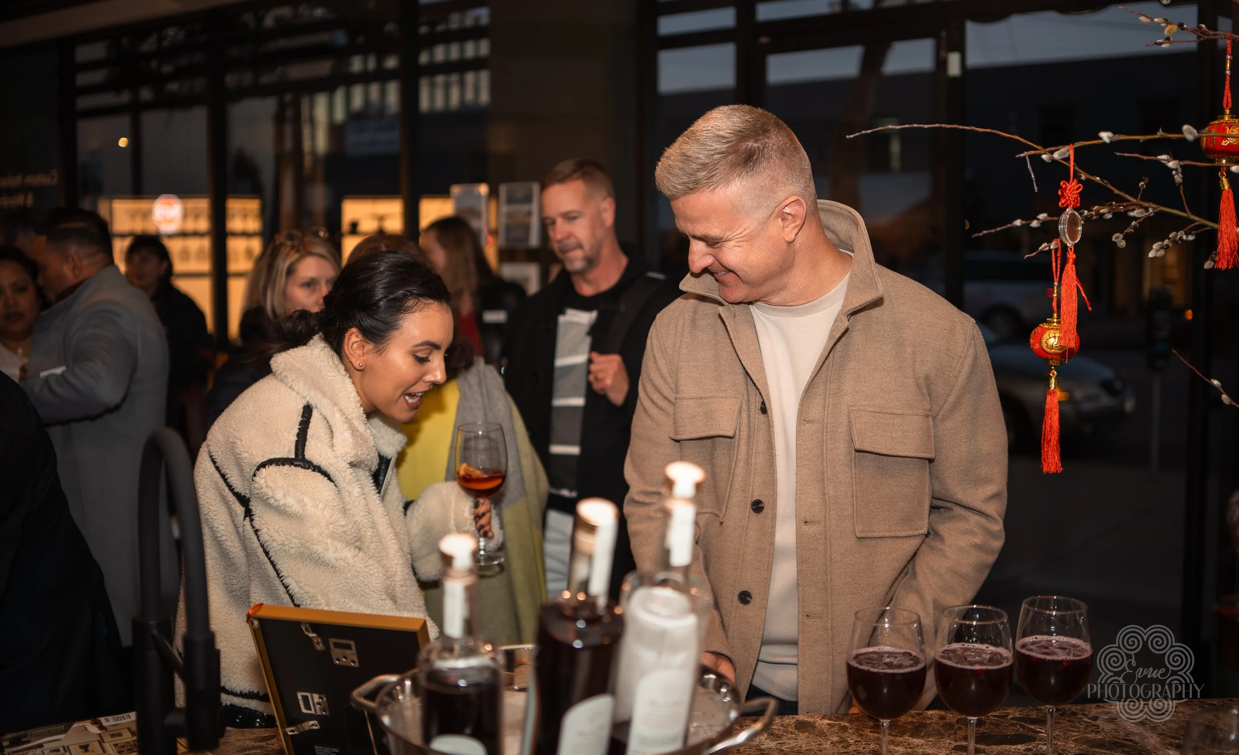 A group of people socializing at an outdoor bar or restaurant during evening, with two individuals in the foreground smiling and talking near a table with three glasses of dark beverages and bottles.