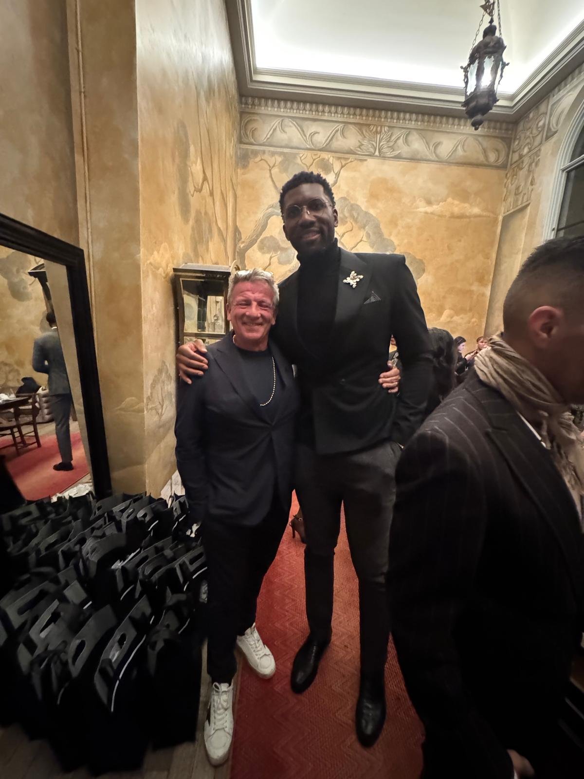 Two men smiling and posing for a photo at an indoor event. One man is shorter with gray hair, wearing a dark suit and white sneakers. The other man is taller, with dark hair and beard, wearing a black suit and tie, black shoes, and a butterfly pin on