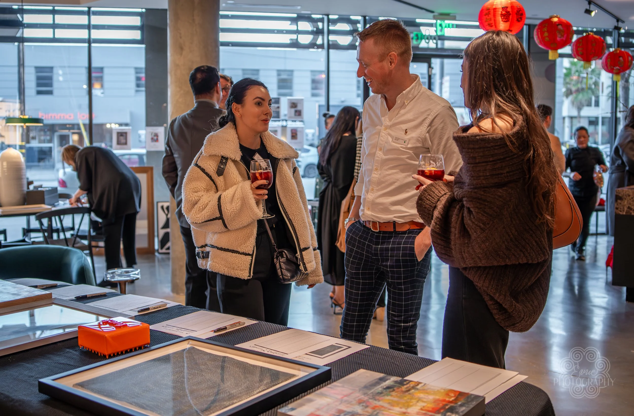 Three people are talking and holding drinks at an indoor event with red lanterns hanging overhead and large windows in the background.