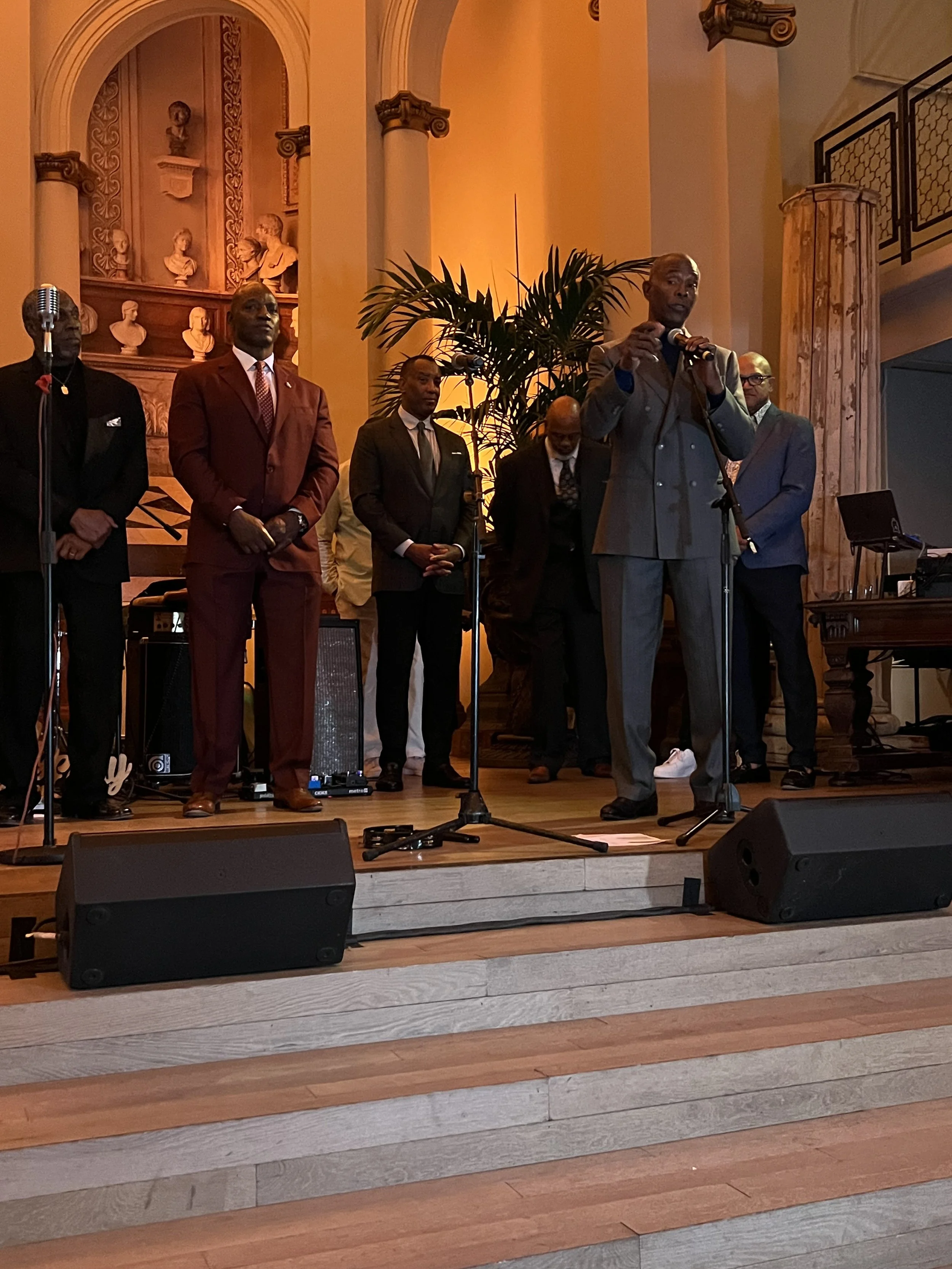 A group of men dressed in formal suits standing on a stage with a man speaking into a microphone. There are musical equipment and a large potted plant behind them in a grand, ornate room with tall columns and bust sculptures in the background.