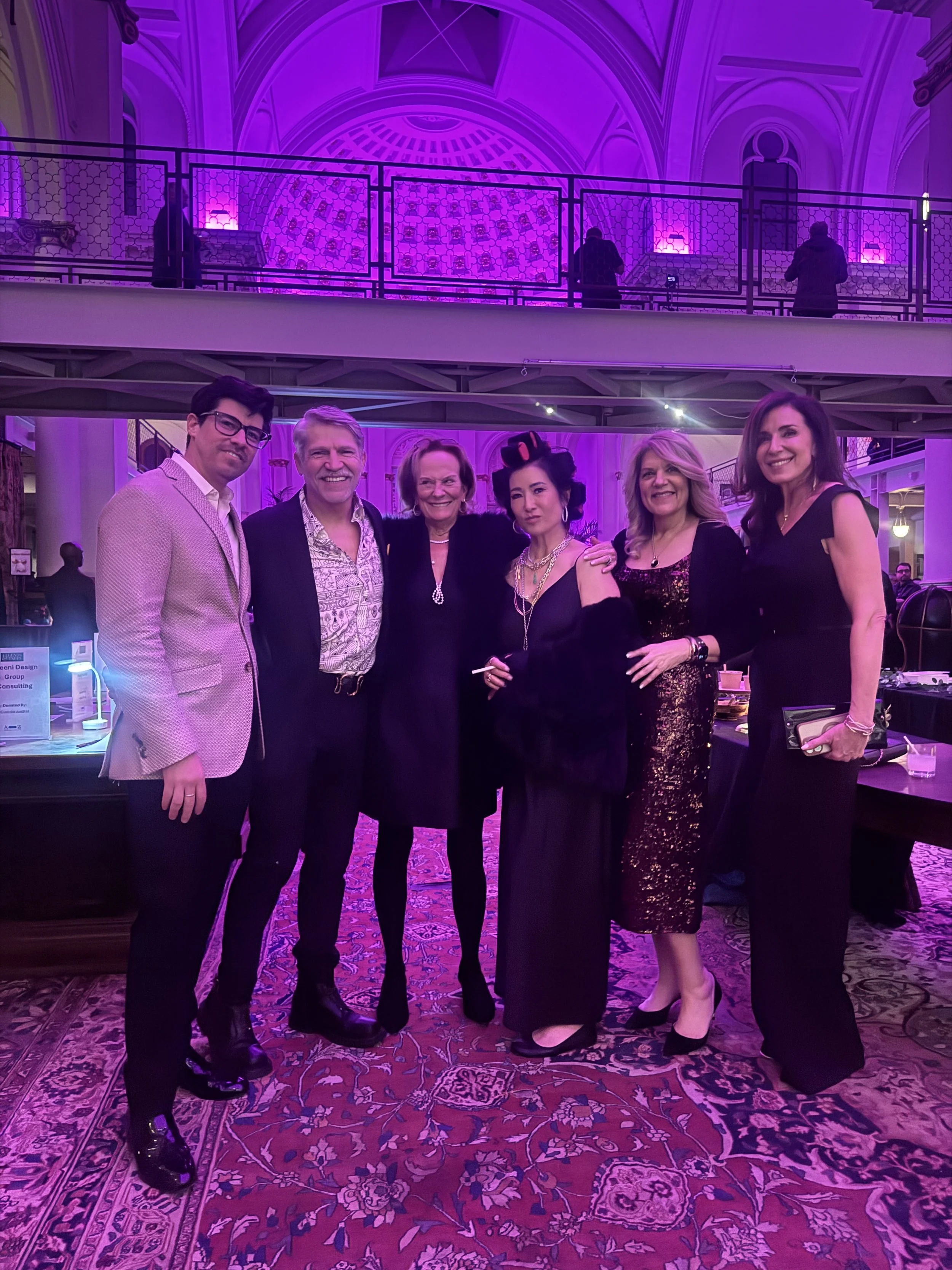 A group of six people dressed in formal attire posing together inside a venue with purple lighting and ornate architecture.