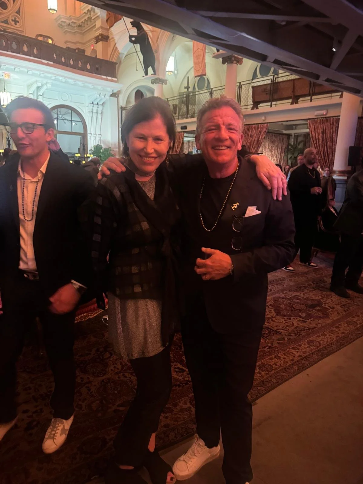 Two smiling people, a woman and a man, are posing together indoors at a lively event with a decorative interior. The woman has short dark hair and is wearing a dark jacket over a patterned outfit. The man has short gray hair and is dressed in black, 