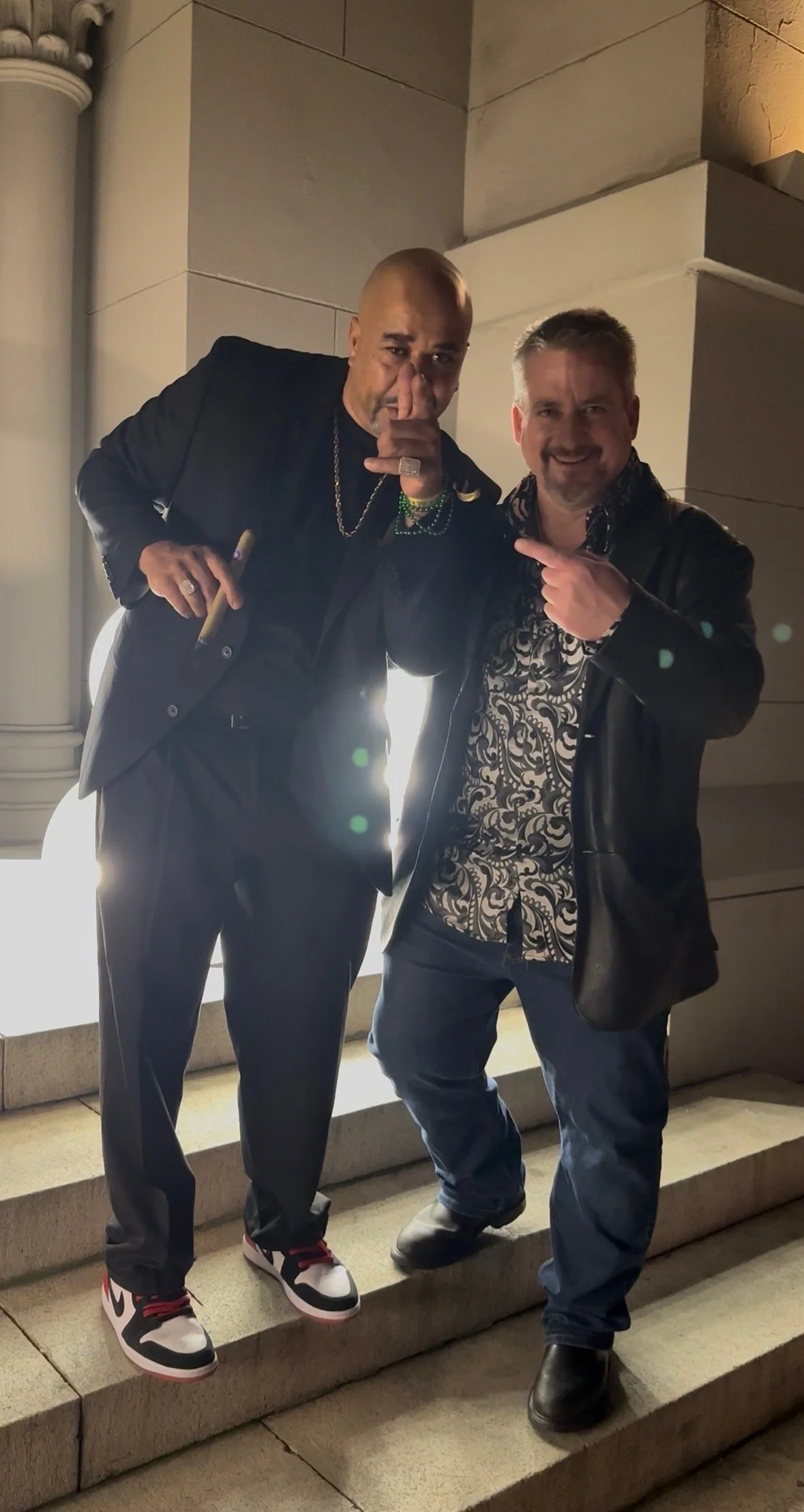 Two men standing on stairs at night, one holding a cigar, both smiling and pointing at each other, illuminated by bright backlight.
