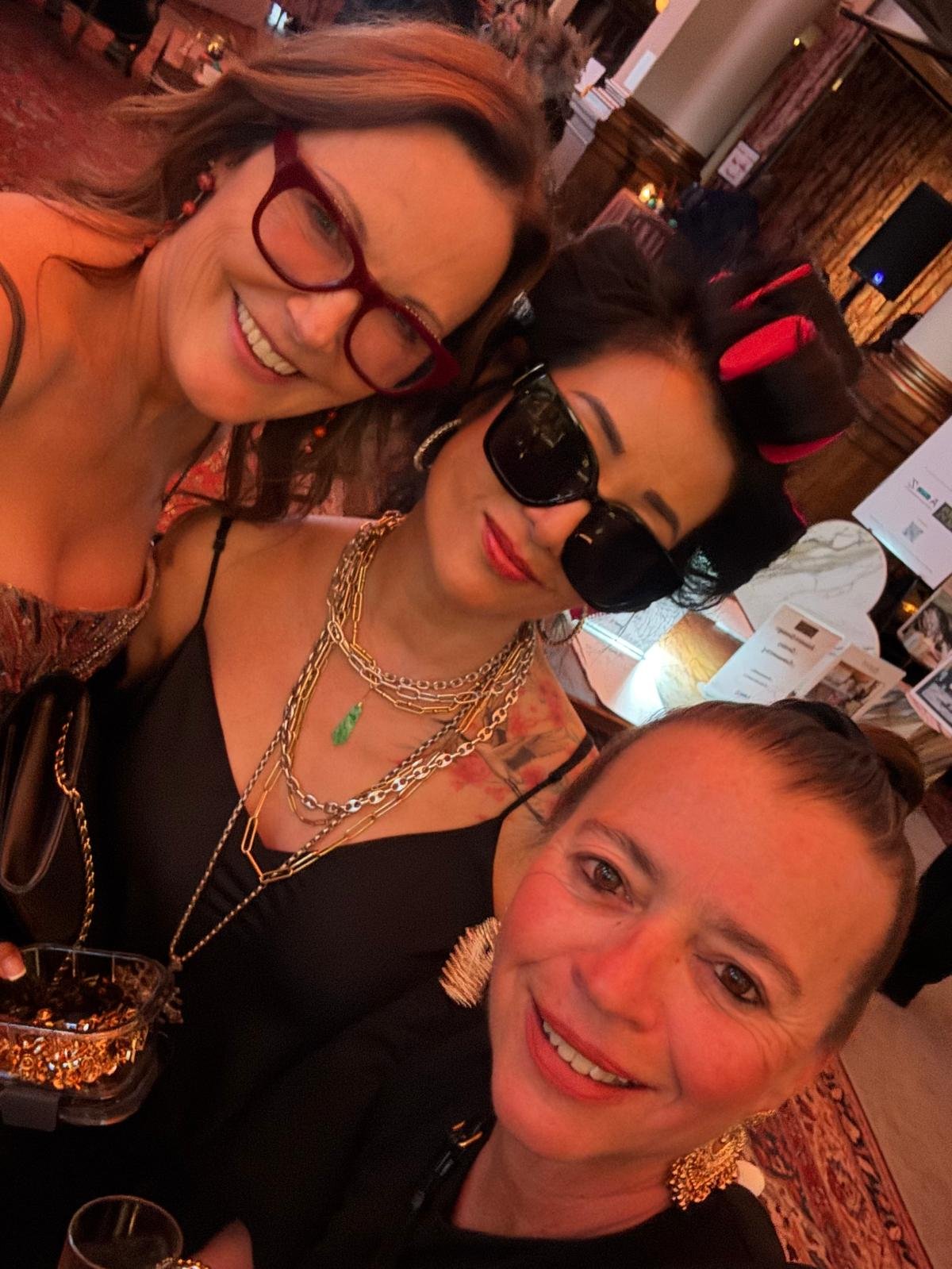 Three people smiling, taking a selfie in a dimly lit restaurant or bar with wooden decor and tables in the background.