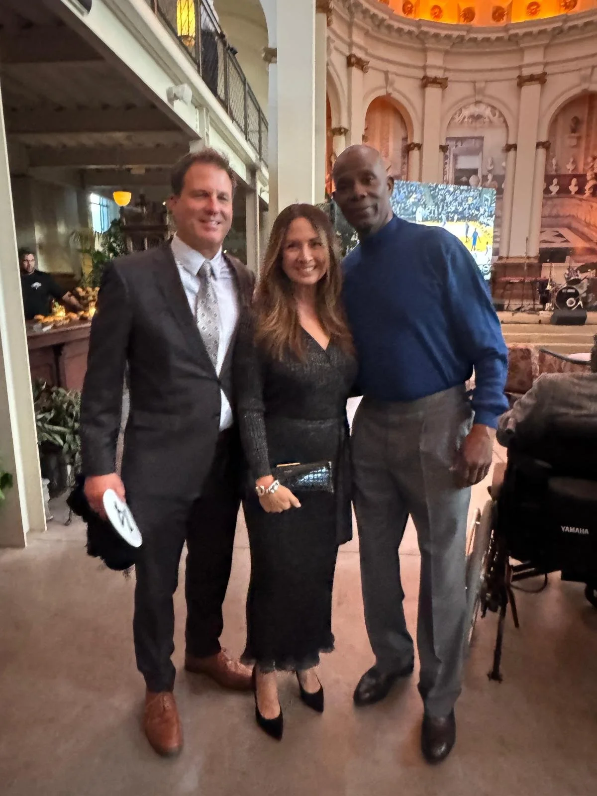 Three people dressed in formal attire, posing inside a decorated venue. The man on the left is wearing a dark suit, white shirt, and a patterned tie. The woman in the middle is wearing a black dress, holding a black clutch and wearing black heels. Th