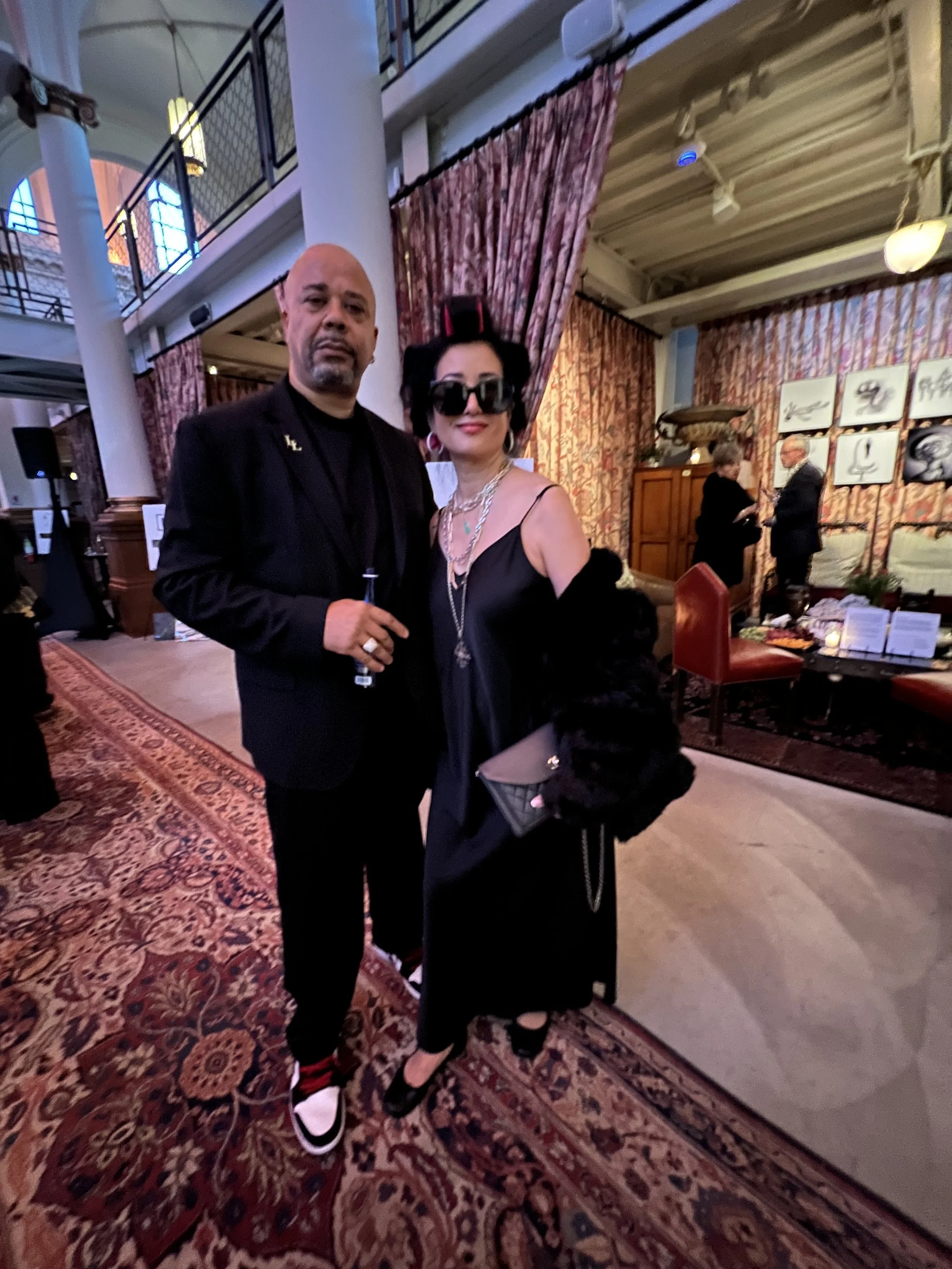 A man and a woman dressed in gothic attire posing indoors at a formal event with ornate curtains, art pieces, and people conversing in the background.