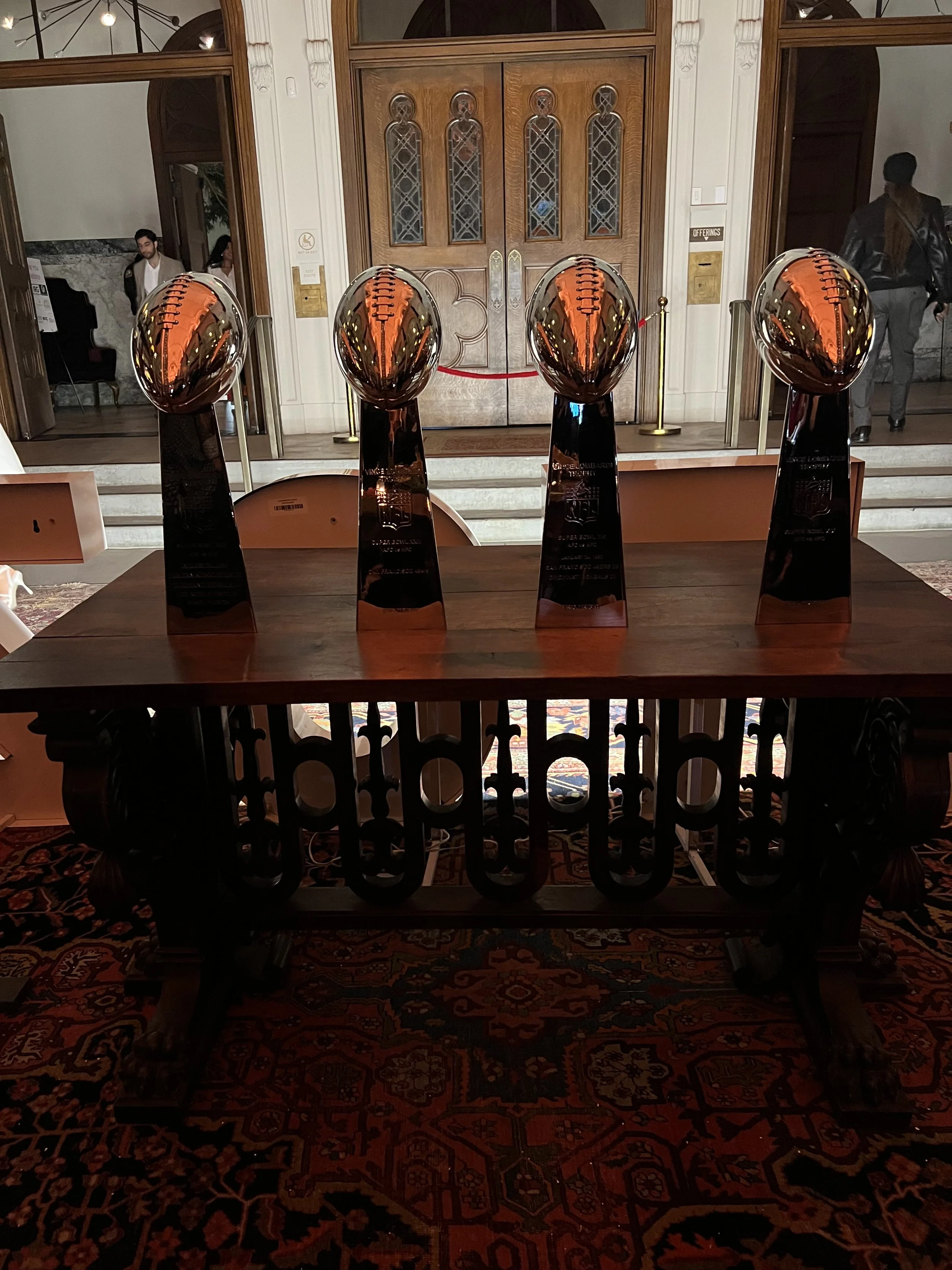 Four Vince Lombardi trophies displayed on a wooden table in a formal room, with large wooden doors and a red carpet underneath.