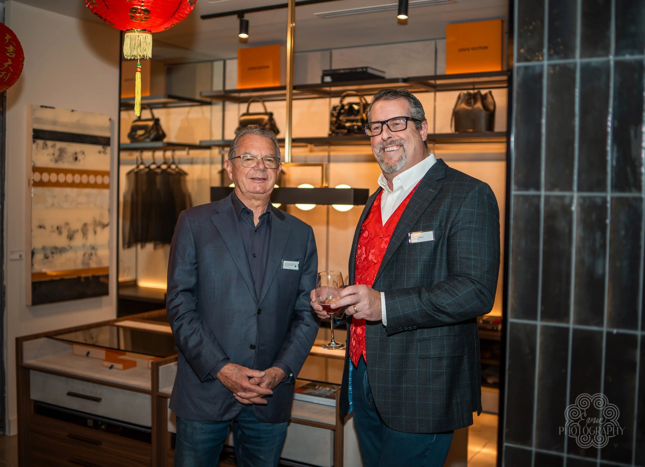 Two men dressed in formal business attire at an indoor event, standing and smiling. One holds a glass of wine. Behind them is a modern interior setting with shelves displaying handbags, boxes, and a clothing rack, decorated with a red lantern hanging