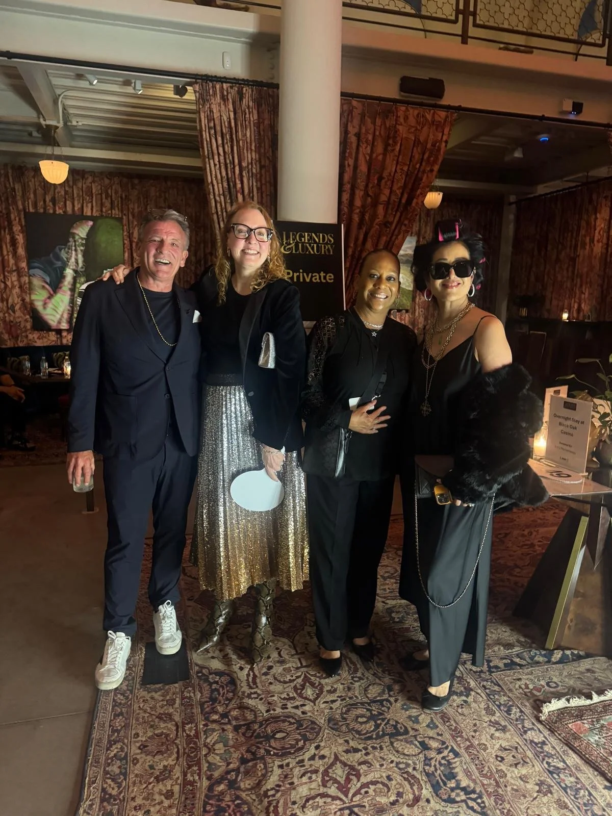 A group of four people standing indoors at an event, smiling for the camera, dressed in formal and fashionable attire. The background features patterned curtains, art pieces on the walls, and a sign that reads 'Legends & Luxury.'