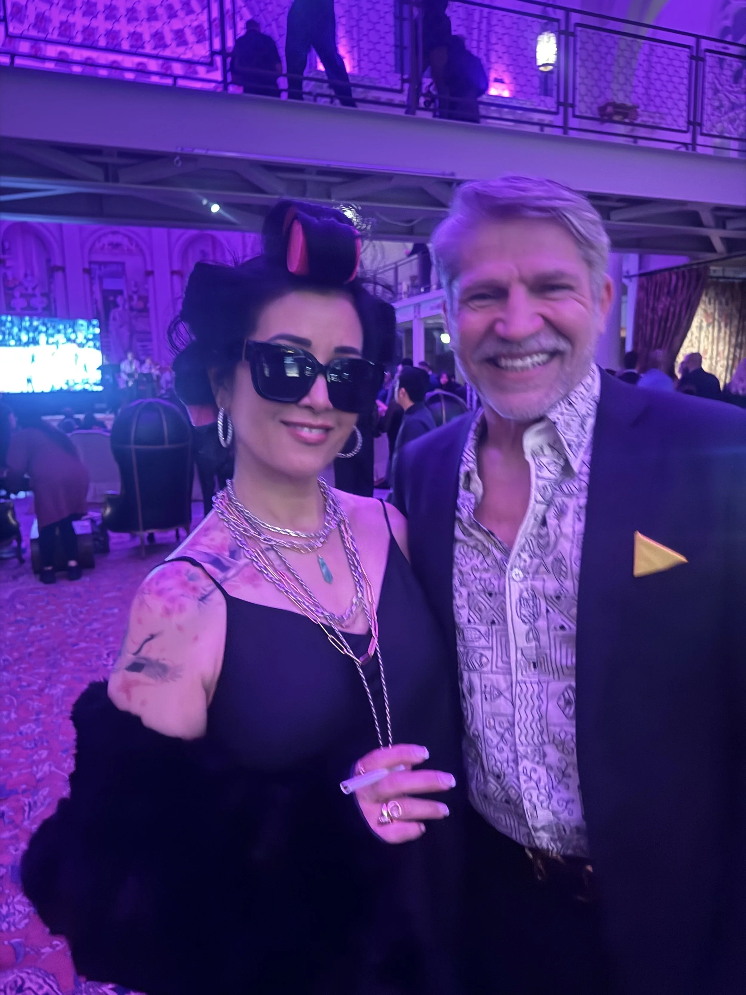 Two people posing for a photo at an event with purple lighting; woman has black hair, sunglasses, jewelry, and tattoos, man has gray hair, beard, and a patterned shirt under a blazer.