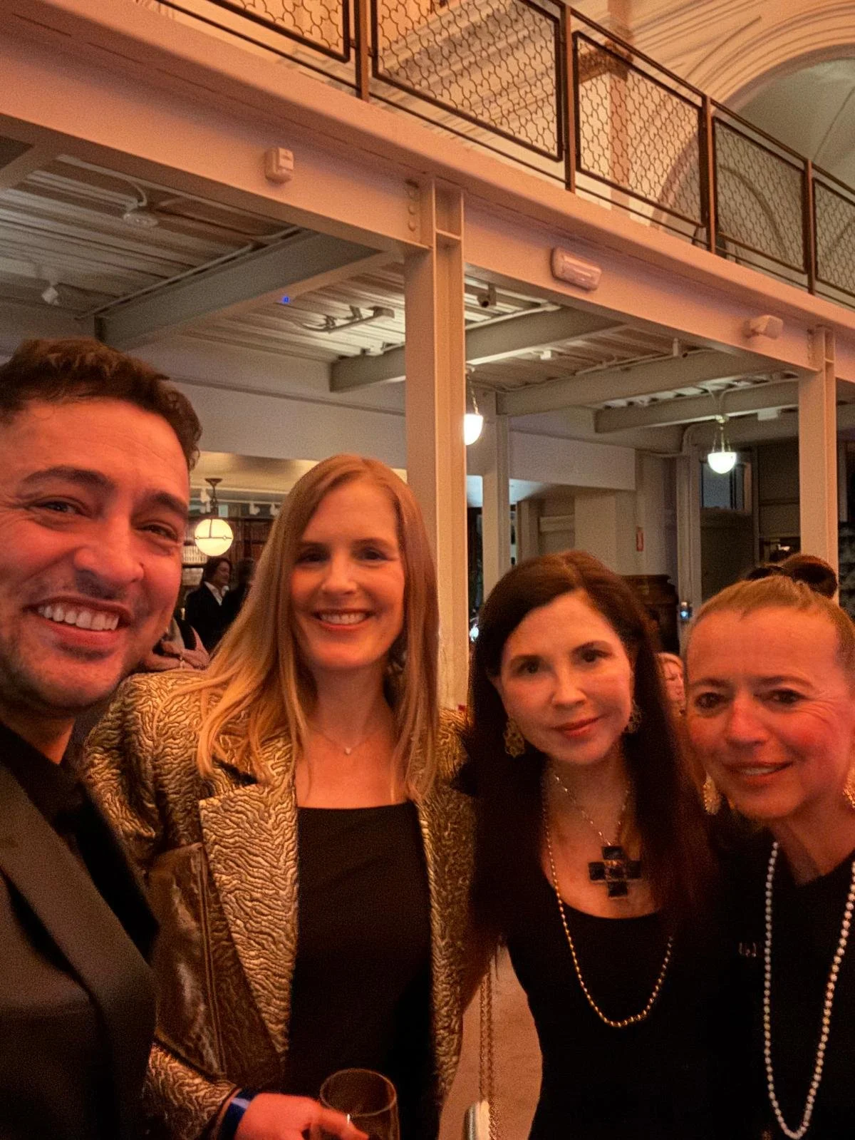 Group of four people smiling and posing together at an indoor event, dressed in formal attire, with warm lighting and decorative interior background.