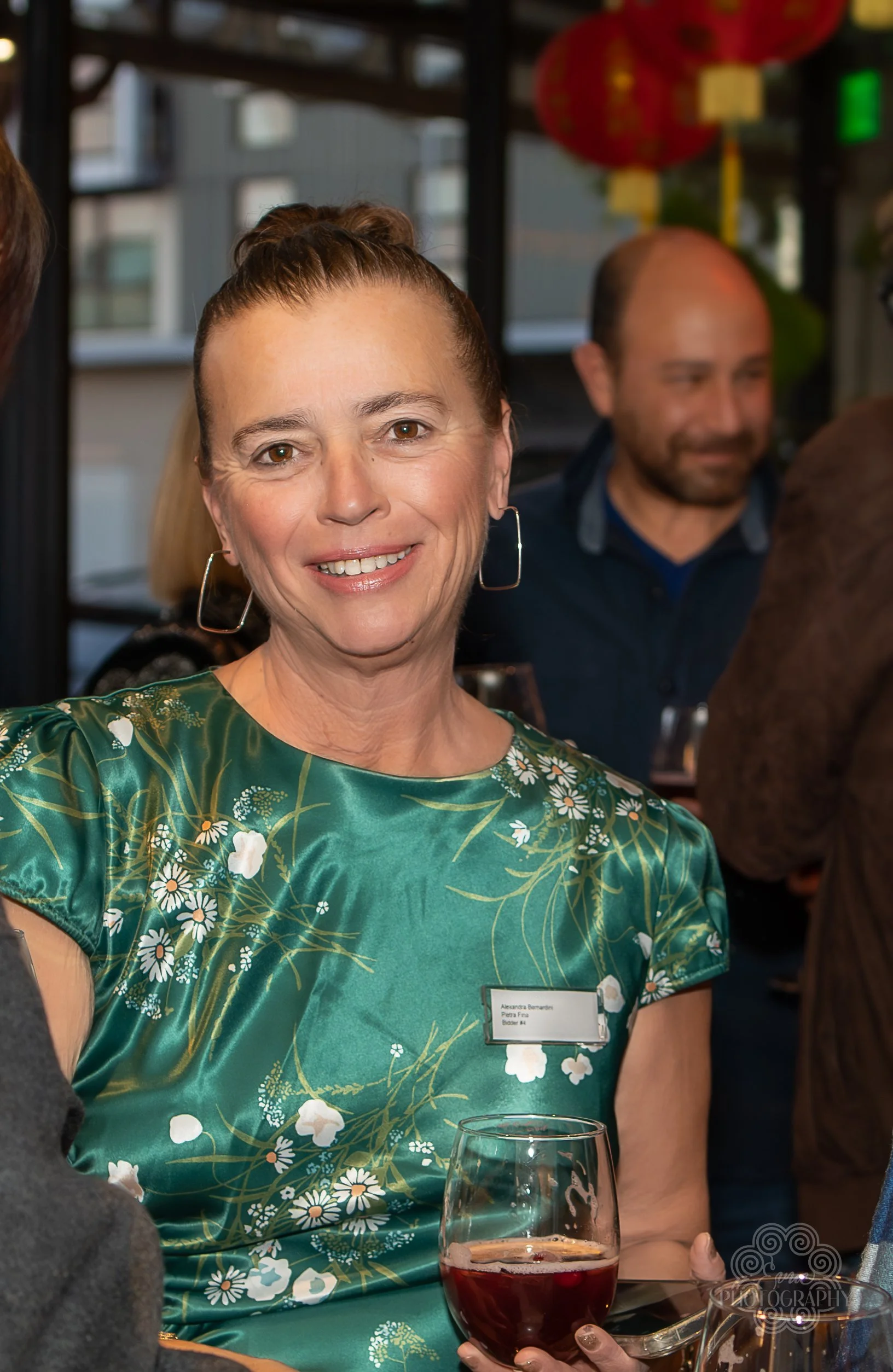 A smiling woman wearing a green floral dress and hoop earrings, holding a glass of red wine, at a social gathering indoors.