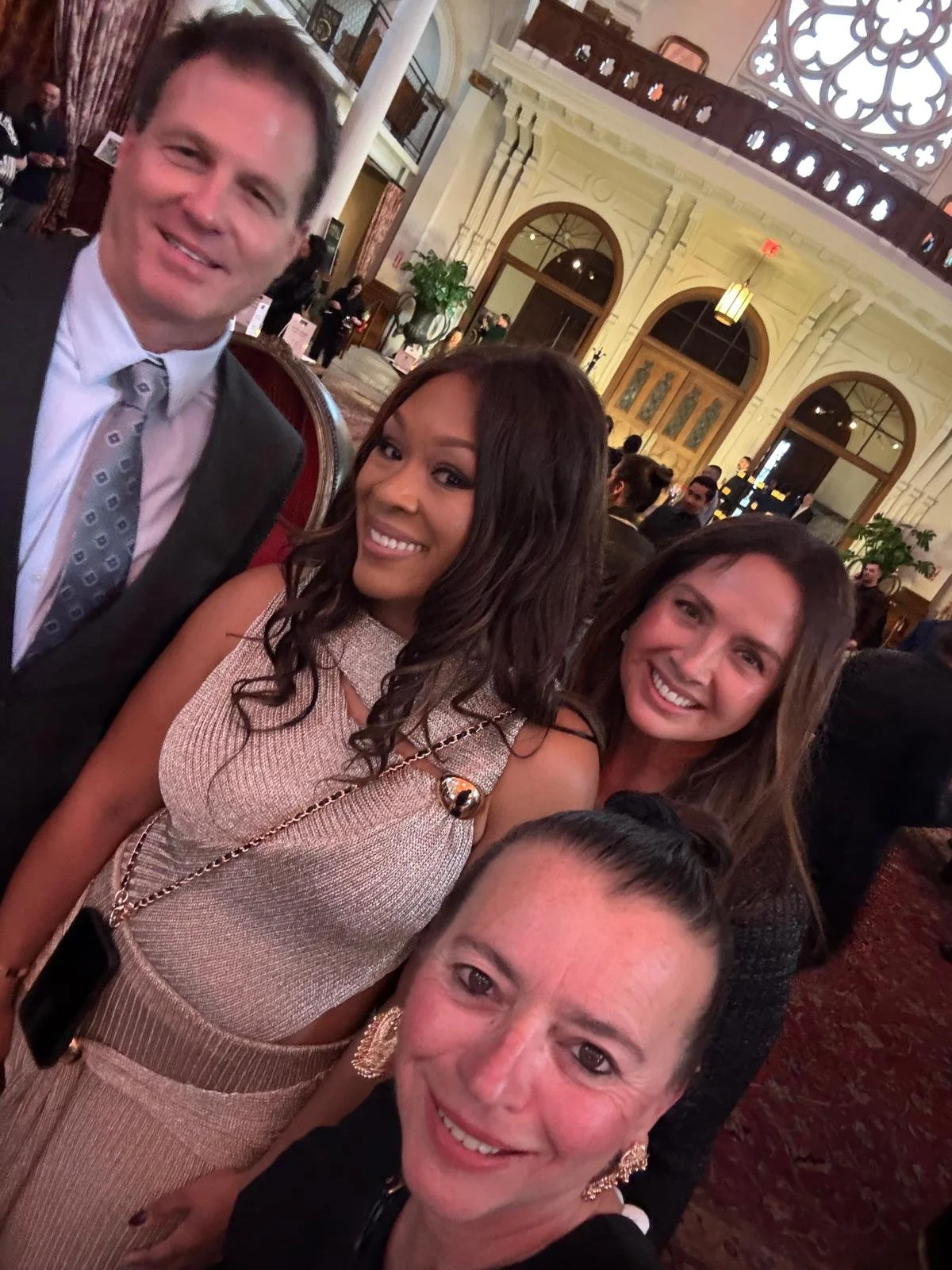 Four people taking a selfie at a formal event inside a grand hall with ornate architecture, large windows, and decorative details.