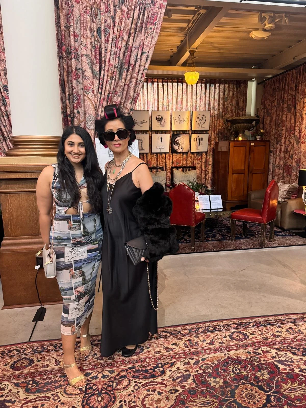 Two women dressed in elegant attire standing in a decorated indoor setting with curtains, artwork, and seating.