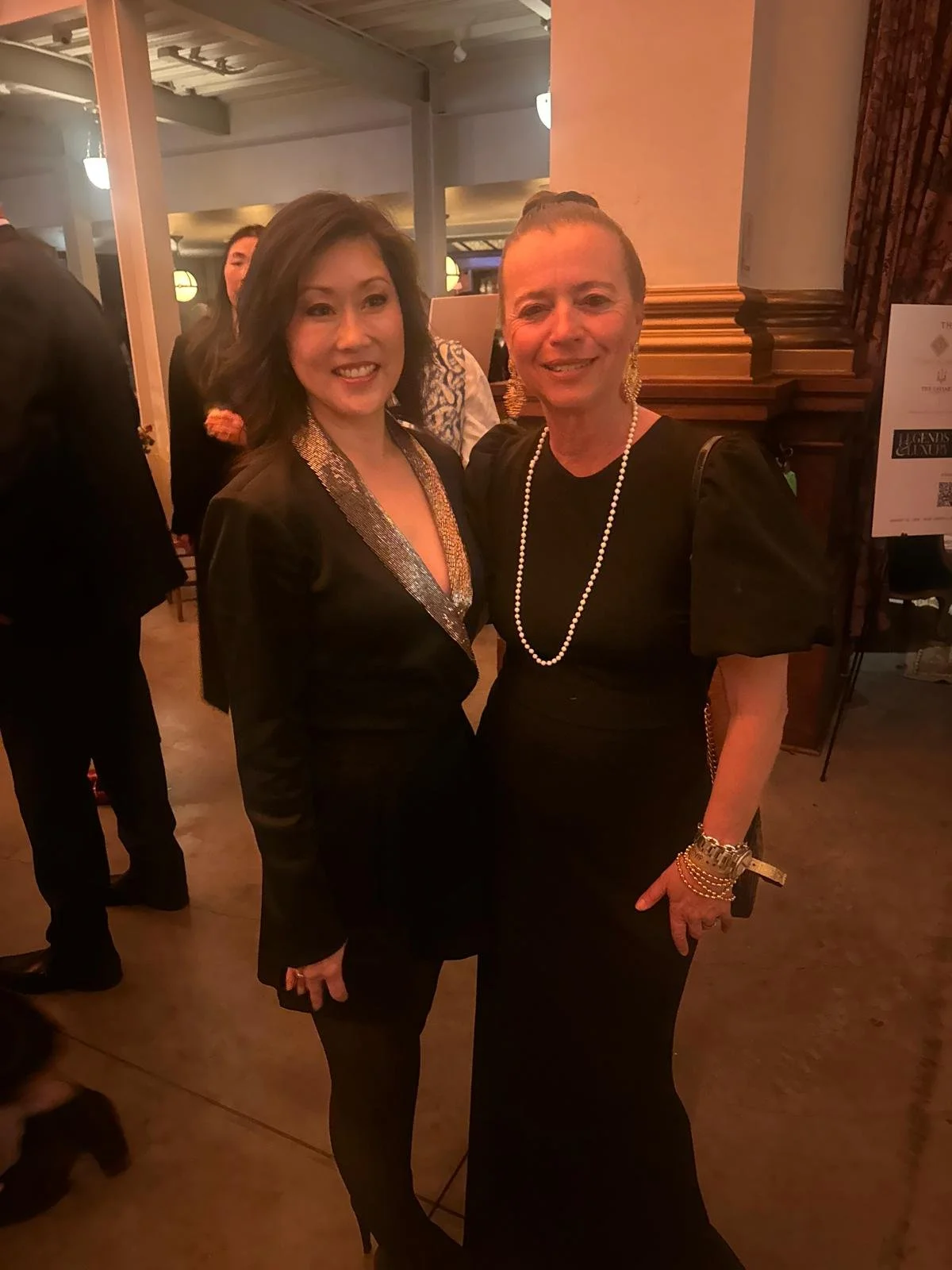 Two women in black dresses smiling at a formal event with warm lighting and other guests in the background.