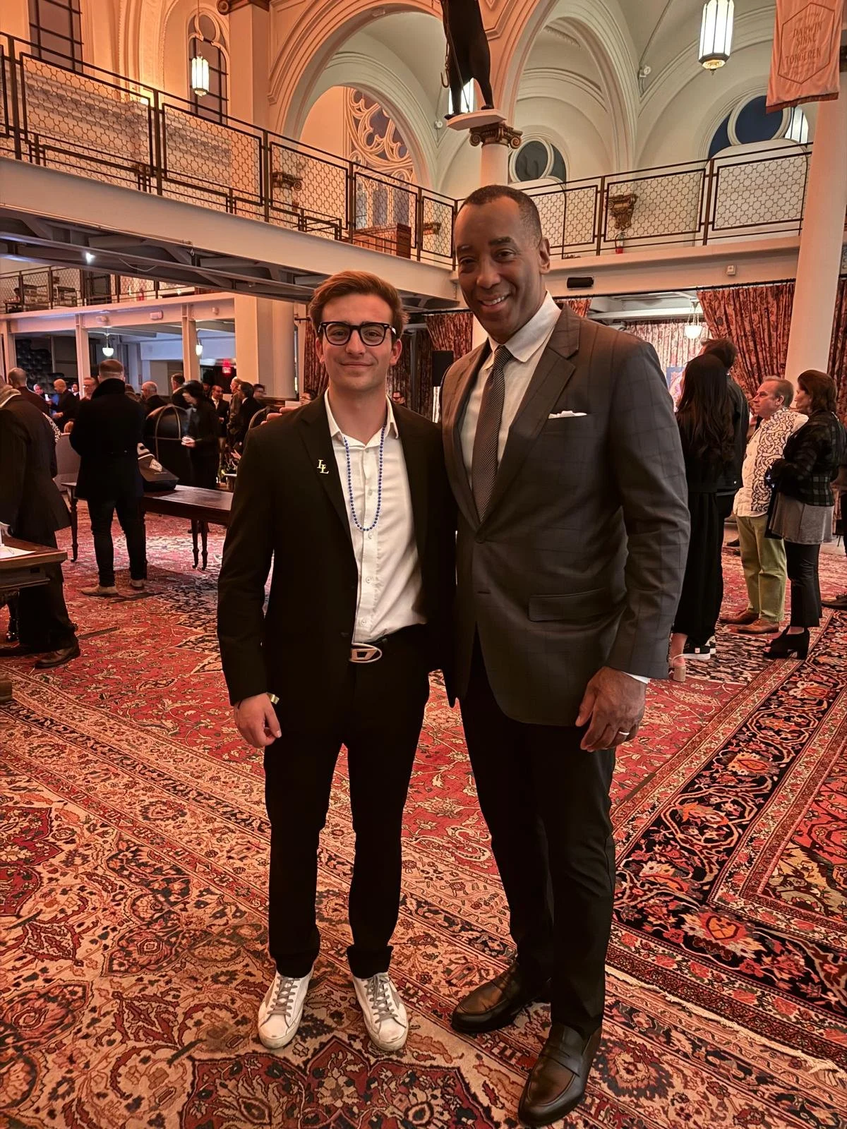 Two men in suits standing together at a formal event inside a large, decorated hall with a patterned rug, high vaulted ceilings, and a balcony. The background shows other attendees mingling.