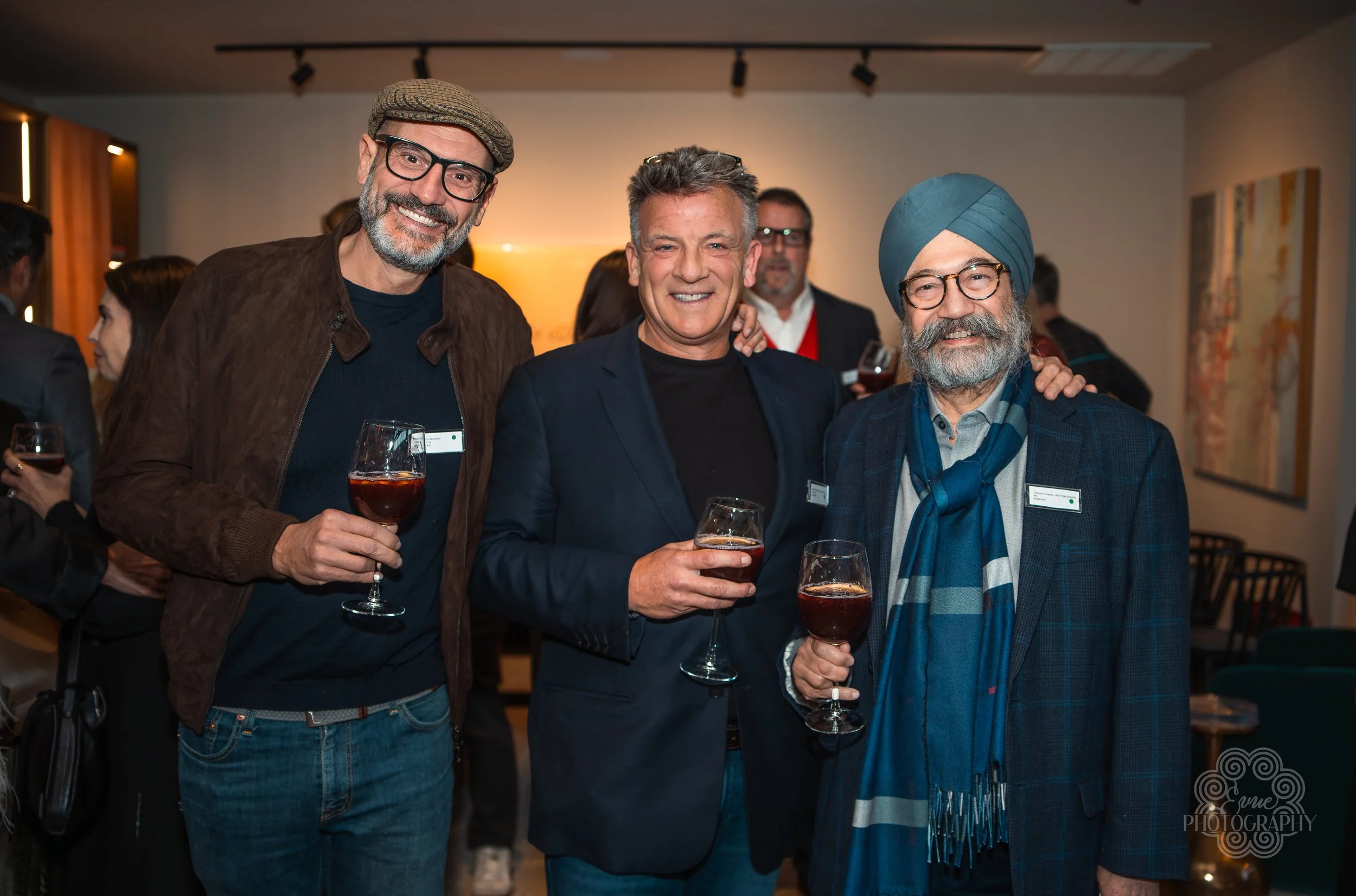 Three men smiling at a social gathering, holding glasses of red wine, with other people and artwork in the background.