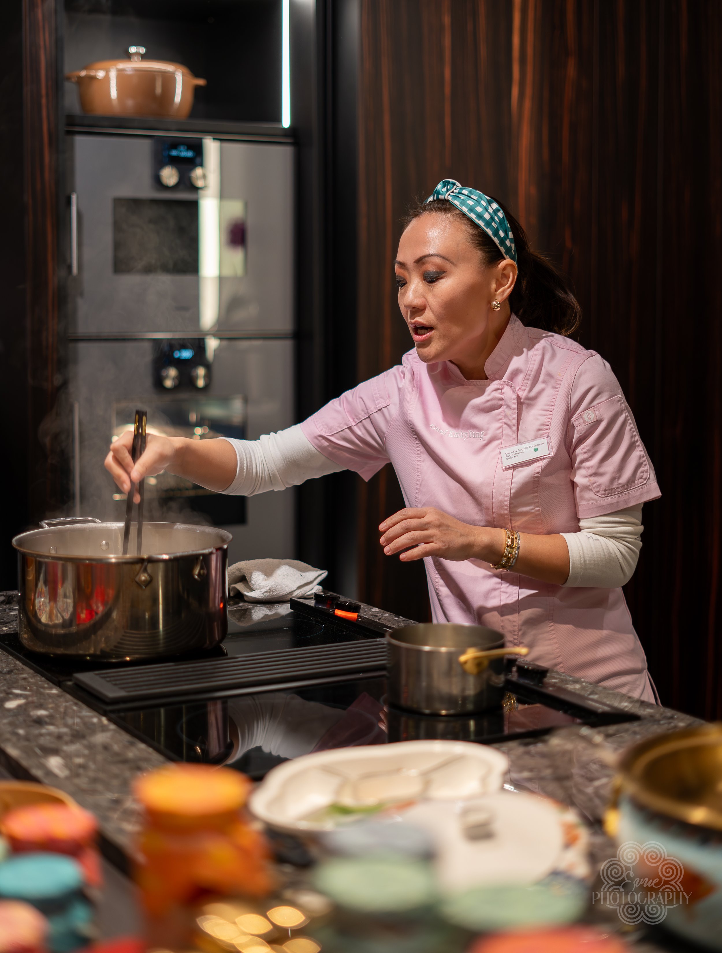 A woman in a pink chef's coat and a headband cooks at a stove in a kitchen with dark wood cabinets, surrounded by colorful decorated cupcakes on the counter.