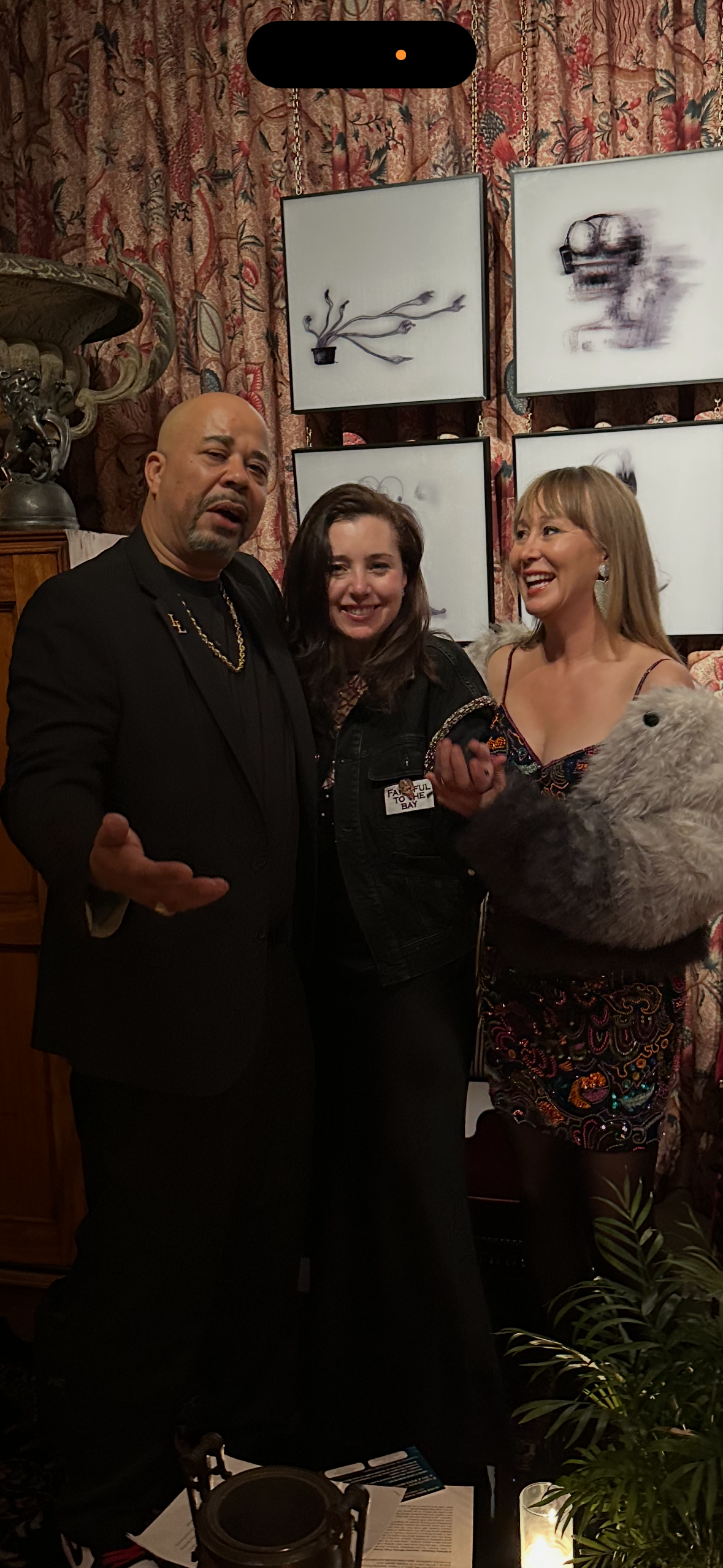 Three people posing together inside a room with patterned curtains and artwork on the wall behind them. One woman is holding a stuffed animal.